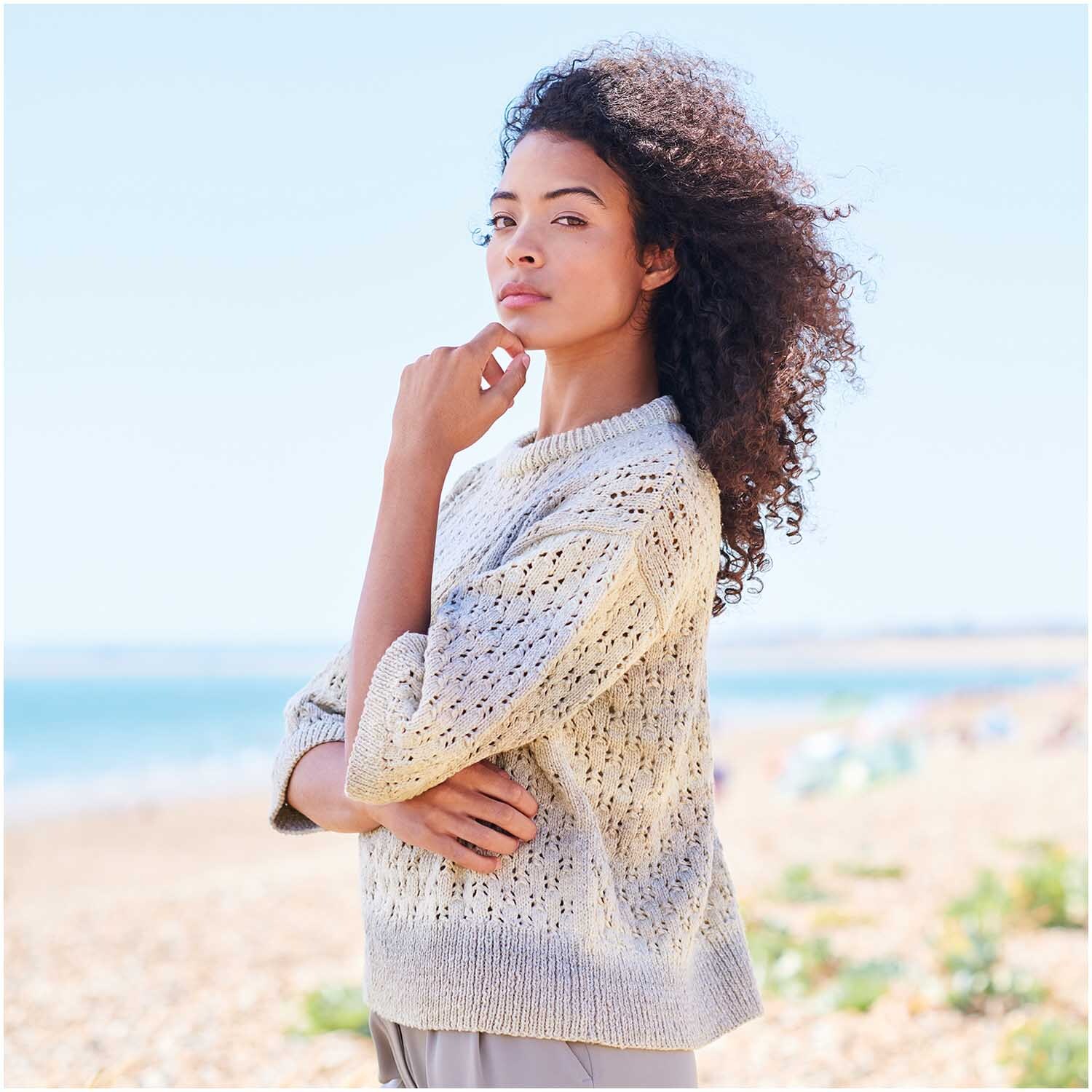 Fashion Cotton Light & Long dk: Frau am Strand in hellem Strickpullover, windiges Haar, Meereshintergrund.