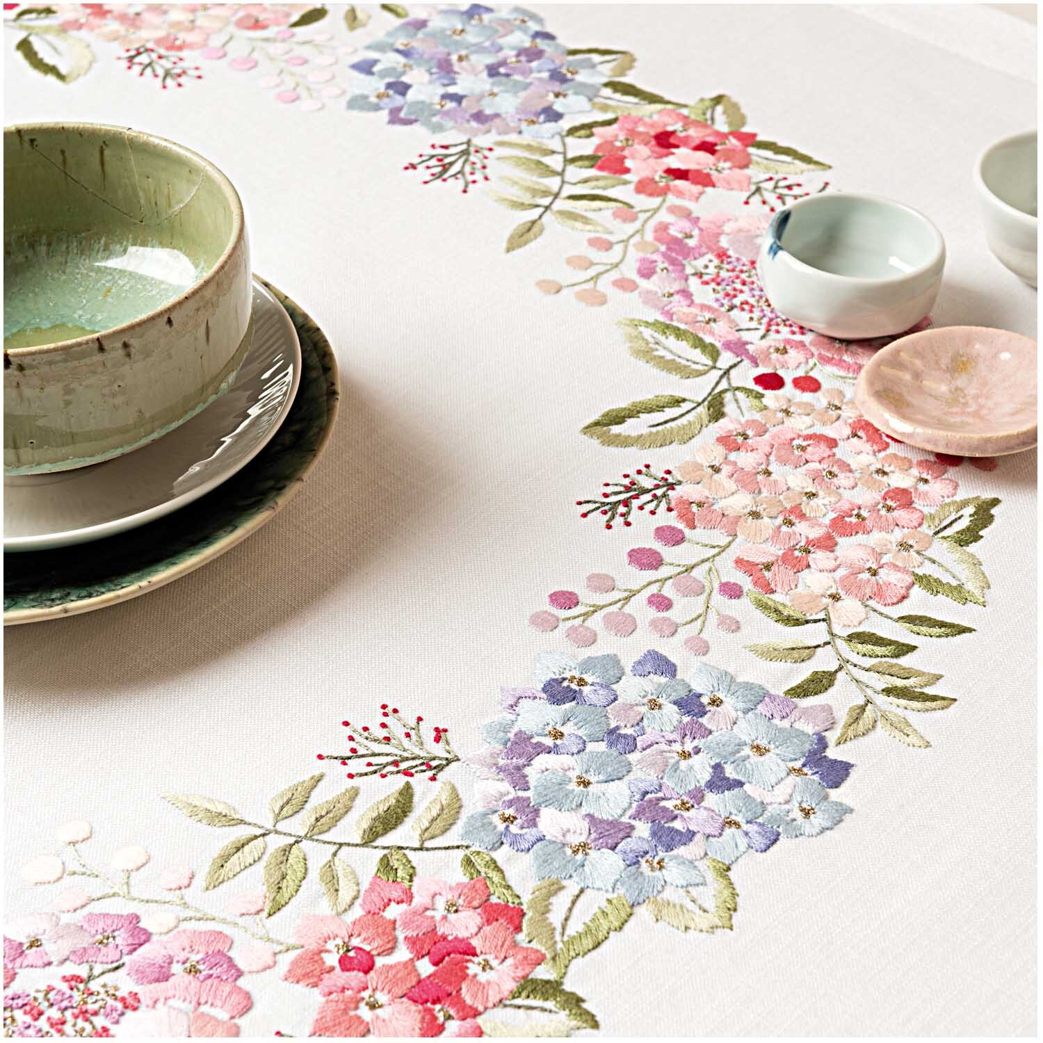 Stickpackung Tischdecke Hortensien 90x90cm mit floralen Mustern in Rosa und Blau, umgeben von Geschirr. Marke: Rico Design.