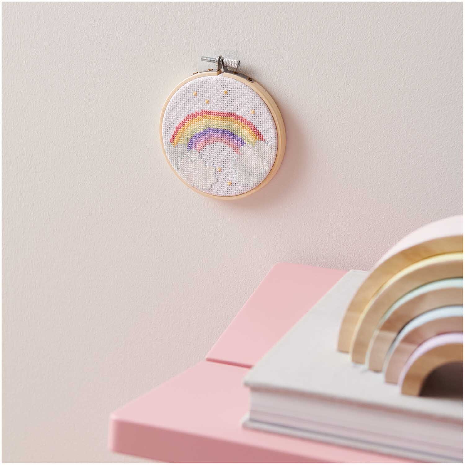 Stickpackung Mini Kit Regenbogen Bild Ø10 cm zeigt einen bunten Regenbogen auf weißem Stoff, aufgehängt an einer rosa Wand.