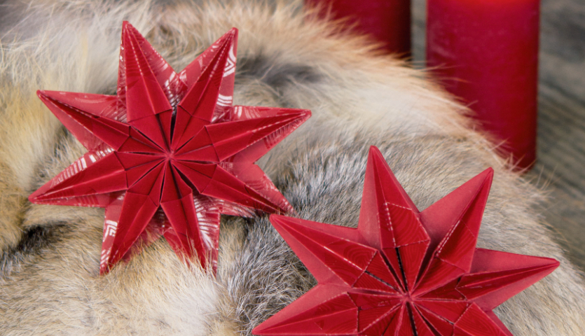 Bastelanleitung Origami-Stern falten: Zwei rote Origami-Sterne auf weichem Fell, im Hintergrund rote Kerzen.