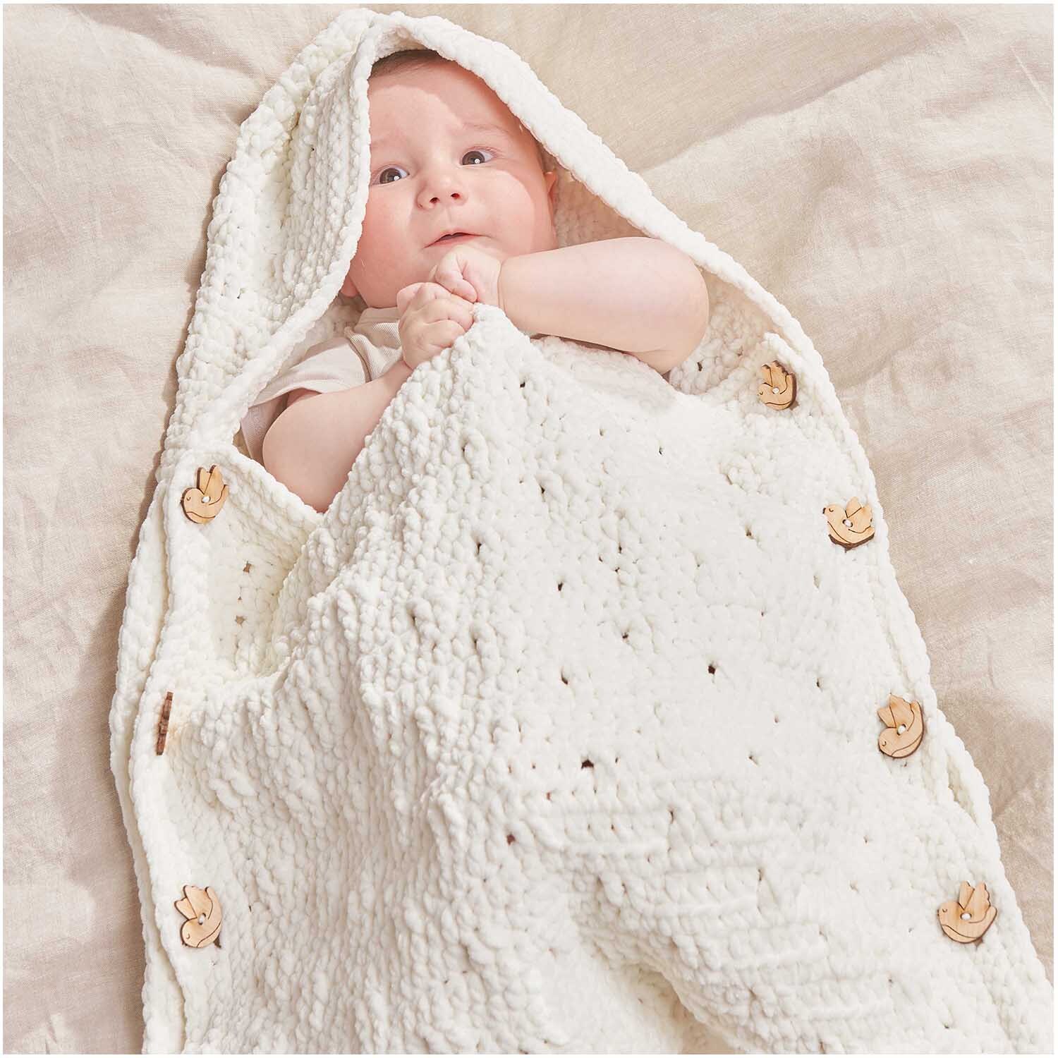 Creative Chenillove: Baby in cremefarbener Decke aus weichem Garn, verziert mit Holzknöpfen. Perfekt für kuschelige Momente.