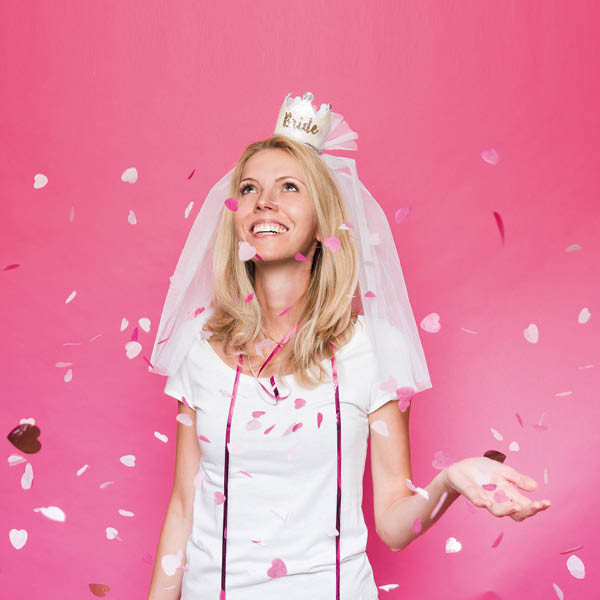 Lachende Frau in weißem T-Shirt und Haarreif mit Krone und Schleier. Fotosetting pinke Wand und Konfetti.
