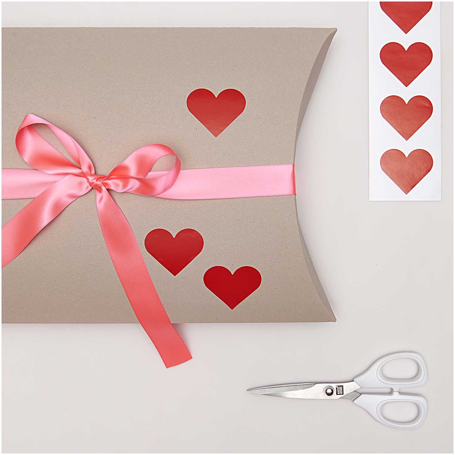 Paper Poetry Sticker Herzen 5cm 120 Stück auf der Rolle. Geschenkverpackung mit roten Herzstickern und rosa Schleife, daneben eine Schere.