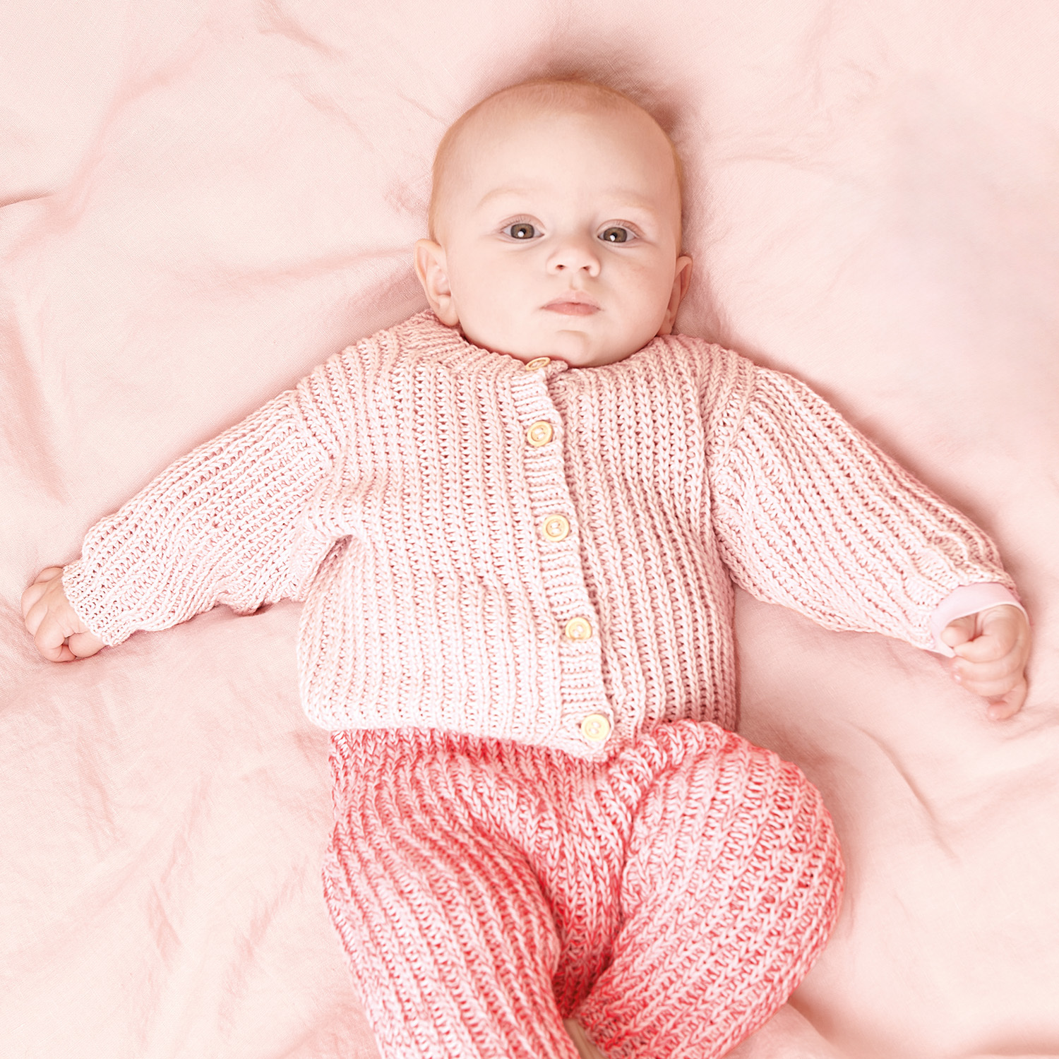 Strickanleitung Jacke aus Rico Baby Cotton Soft dk: Baby in rosa gestrickter Jacke und Hose auf rosa Decke liegend.