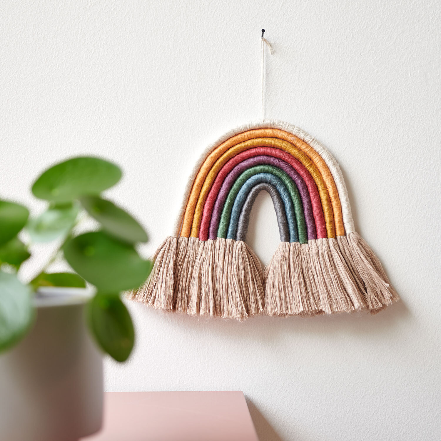 Bastelanleitung Regenbogen mit Sticktwist gestalten: Bunter Wandbehang mit Fransen, dekorativ an weißer Wand.