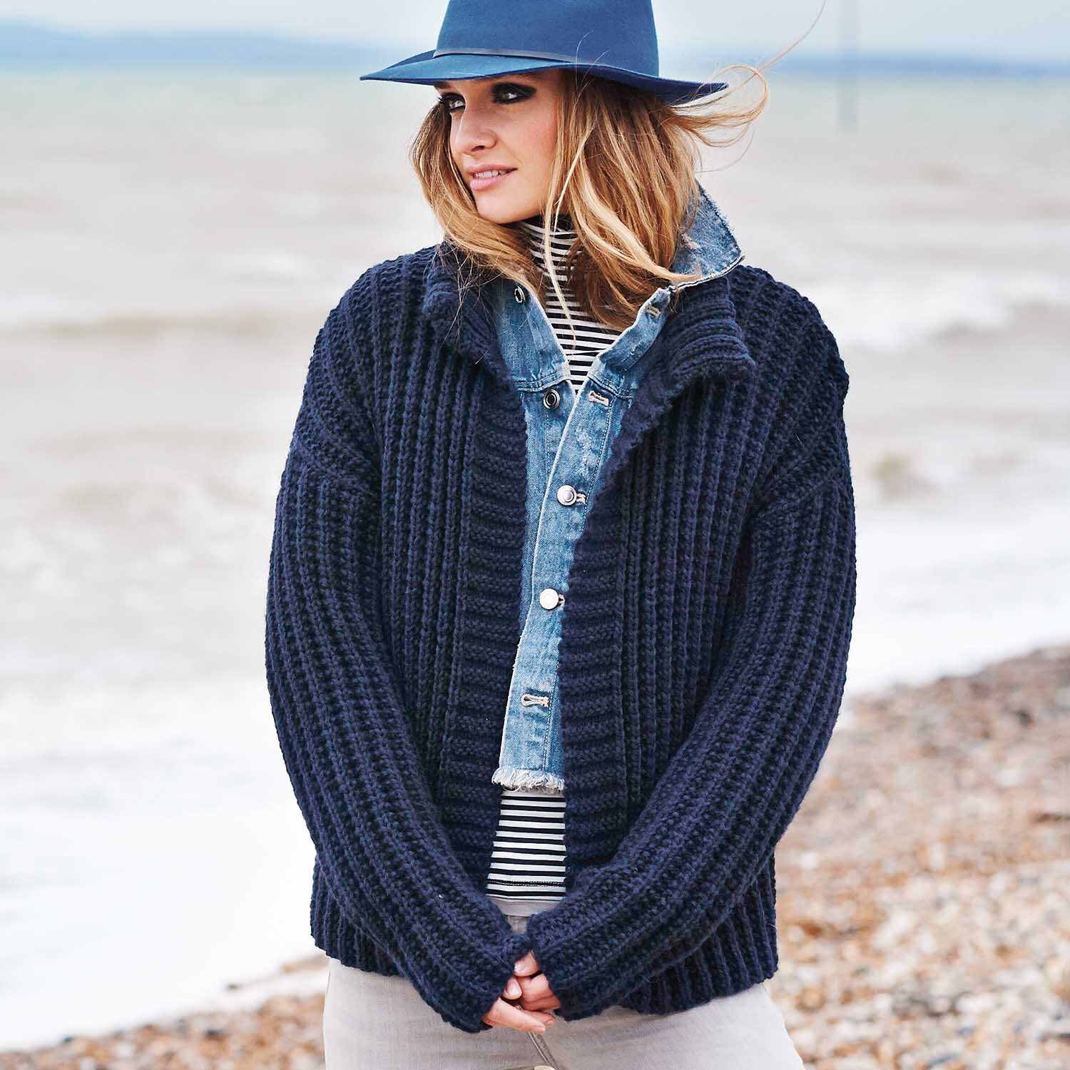 Strickanleitung Rippenmuster-Jacke aus Essentials Alpaca Blend chunky: Frau am Strand in dunkelblauer Strickjacke, gestreiftem Shirt und Jeansjacke.