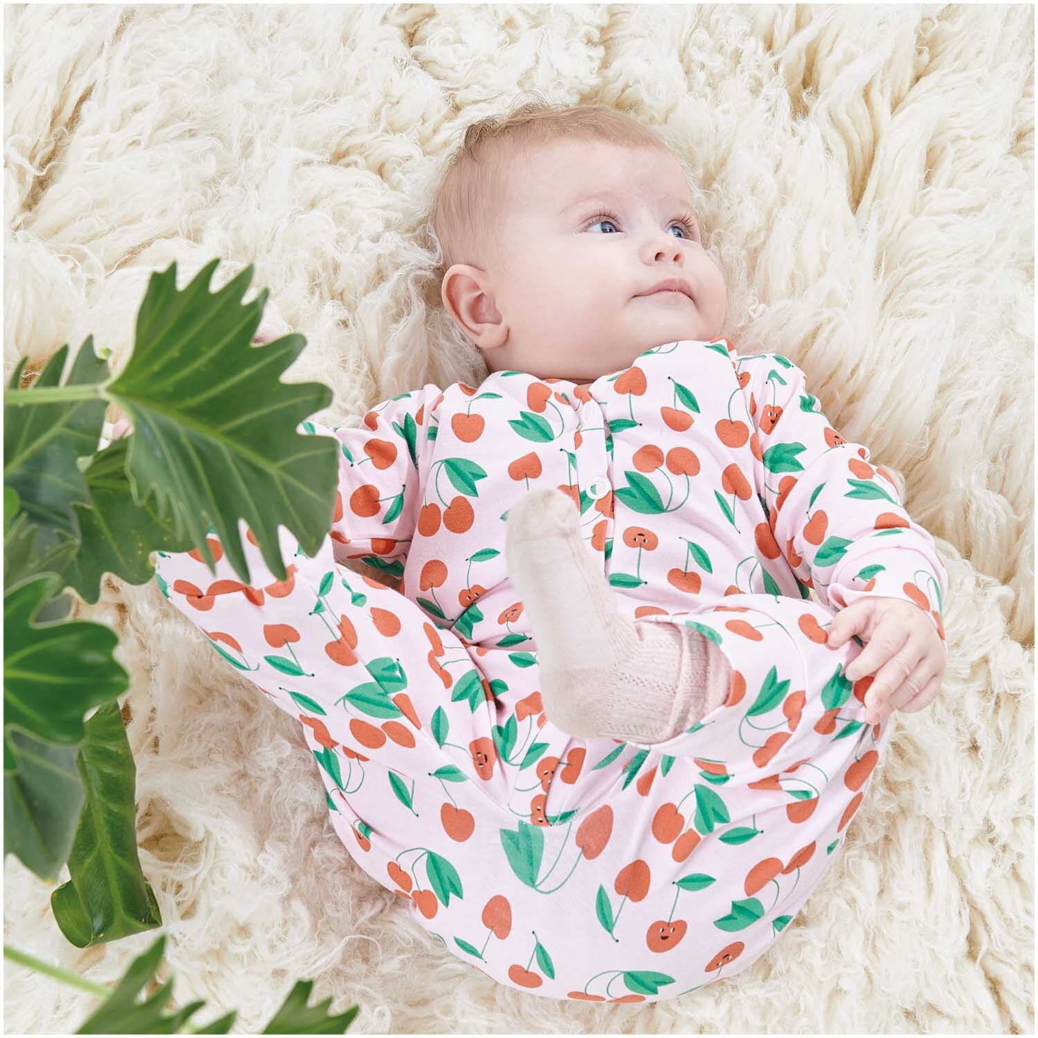 Meterware Jersey bedruckt Kirschen rosa: Ein Baby liegt auf einem flauschigen Teppich, trägt einen rosa Strampler mit Kirschmuster von Rico Design.