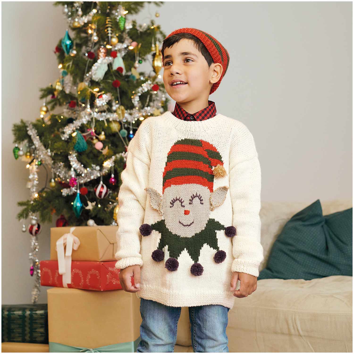 Essentials Merino Plus dk: Kind im cremefarbenen Pullover mit Weihnachtsmotiv vor geschmücktem Baum.