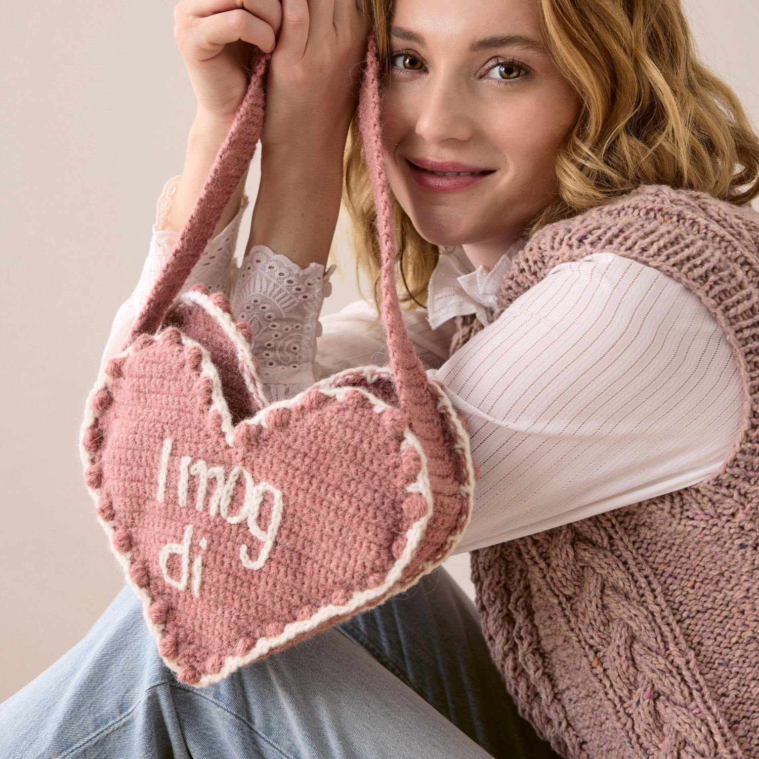 Soft Alpaca Häkelset Herztasche in Rosa, gehalten von einer Frau in einem gestrickten Pullunder.