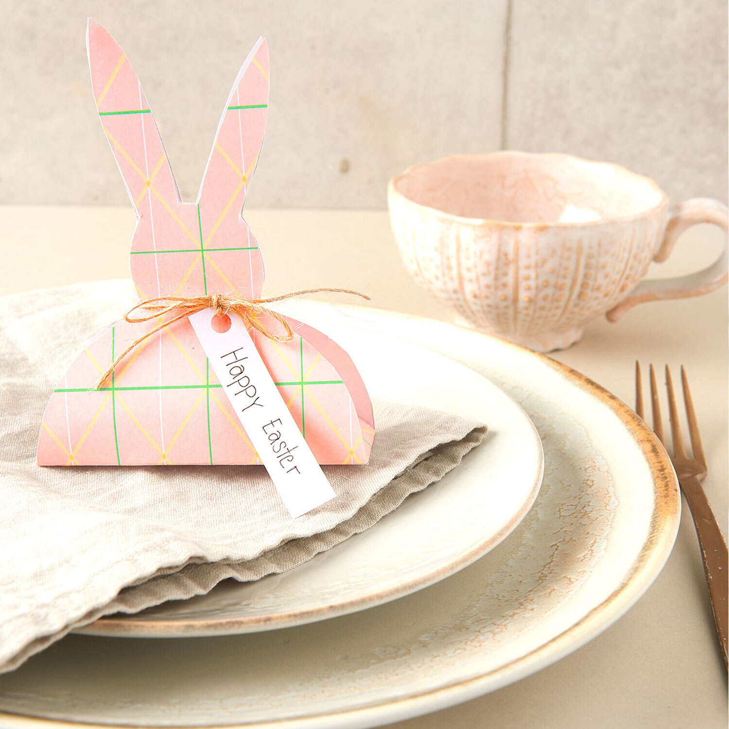 Bastelanleitung Osterhase aus Motivpapier: Ein rosa Papierhase mit Schleife und "Happy Easter"-Schild auf einem gedeckten Tisch.
