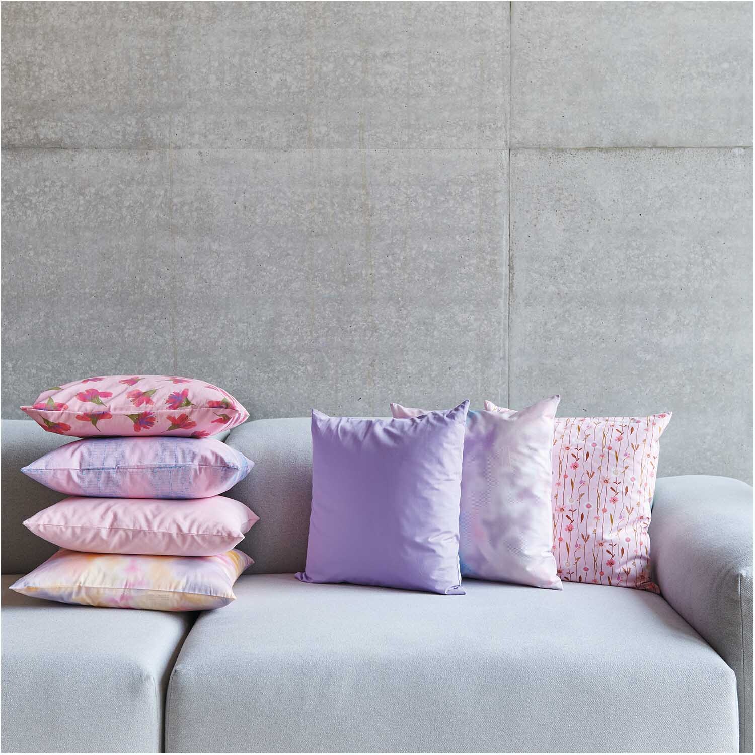 Meterware Druckstoff Blurry Camouflage flieder: Sofakissen in verschiedenen Pastelltönen auf grauem Sofa.