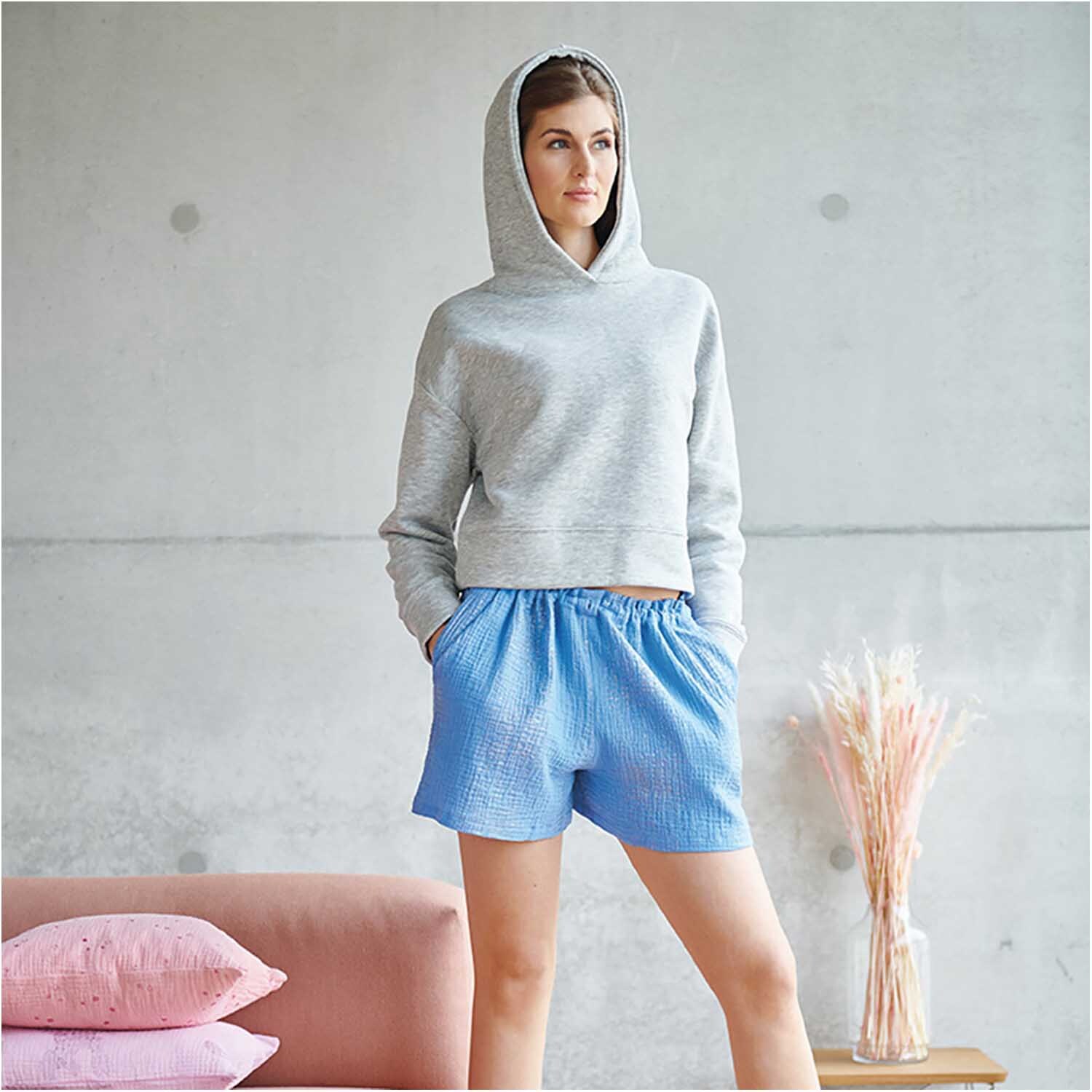 Meterware Krinkelmusselin azur Hot Foil: Frau in grauem Hoodie und blauen Shorts aus Krinkelmusselin, neben rosa Kissen und Trockenblumen.