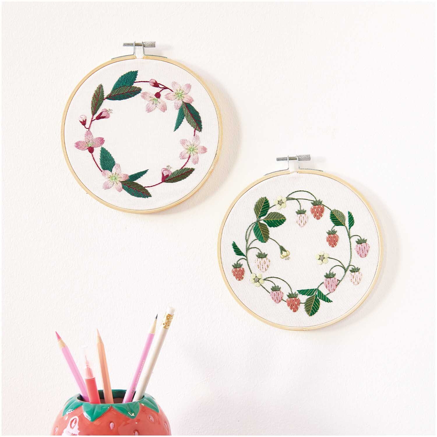 Stickpackung Erdbeerkranz vorgezeichnet: Zwei Stickrahmen mit floralen Motiven, darunter Erdbeeren, an einer weißen Wand. Marke: Rico Design.