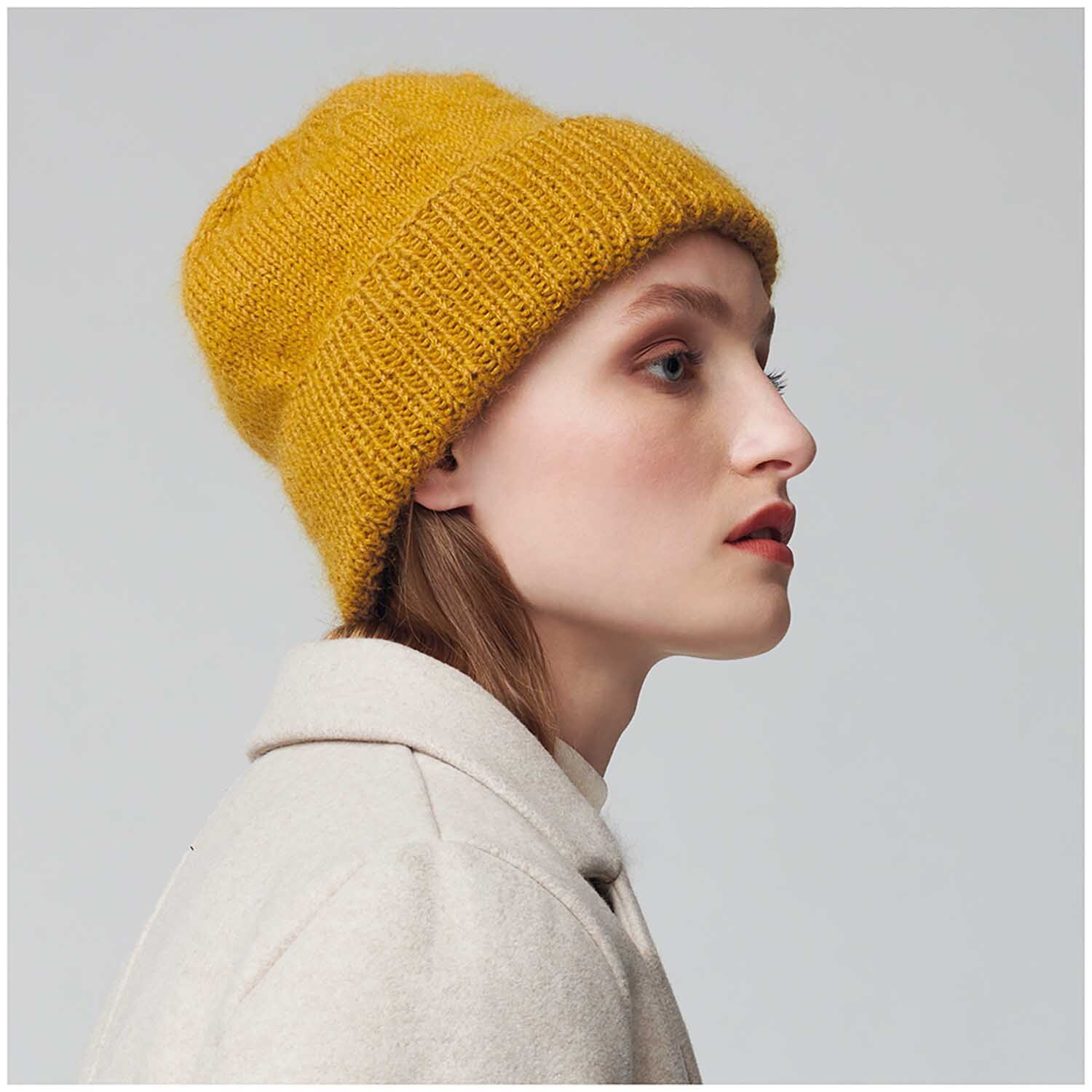 Essentials Merino dk: Frau mit senffarbener Strickmütze aus Rico Design Wolle, seitlich fotografiert.