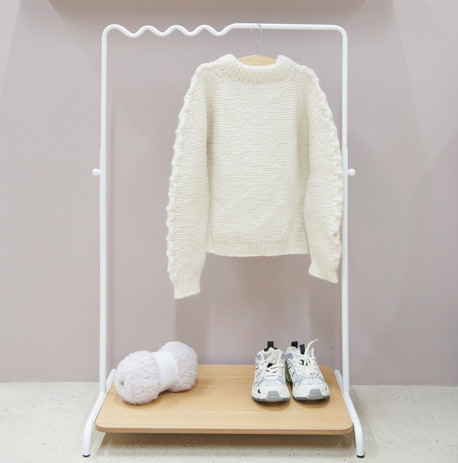 Rico Kids 13 Pullover in Creme auf einem weißen Kleiderständer, daneben weiße Sneaker und ein Knäuel Wolle.