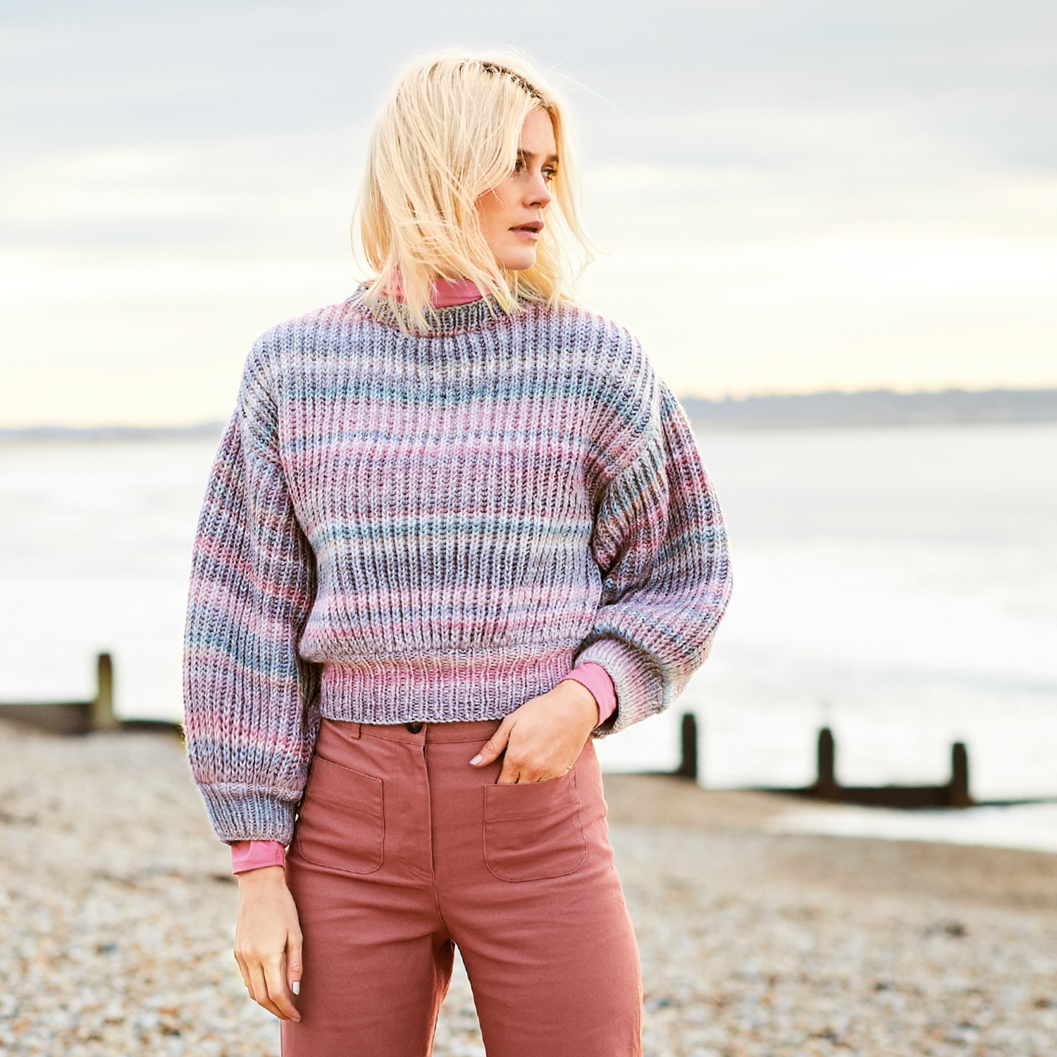 Strickanleitung Pullover aus Creative Melange Chunky: Frau am Strand trägt gestreiften Pullover in Pastelltönen, kombiniert mit rosa Hose.