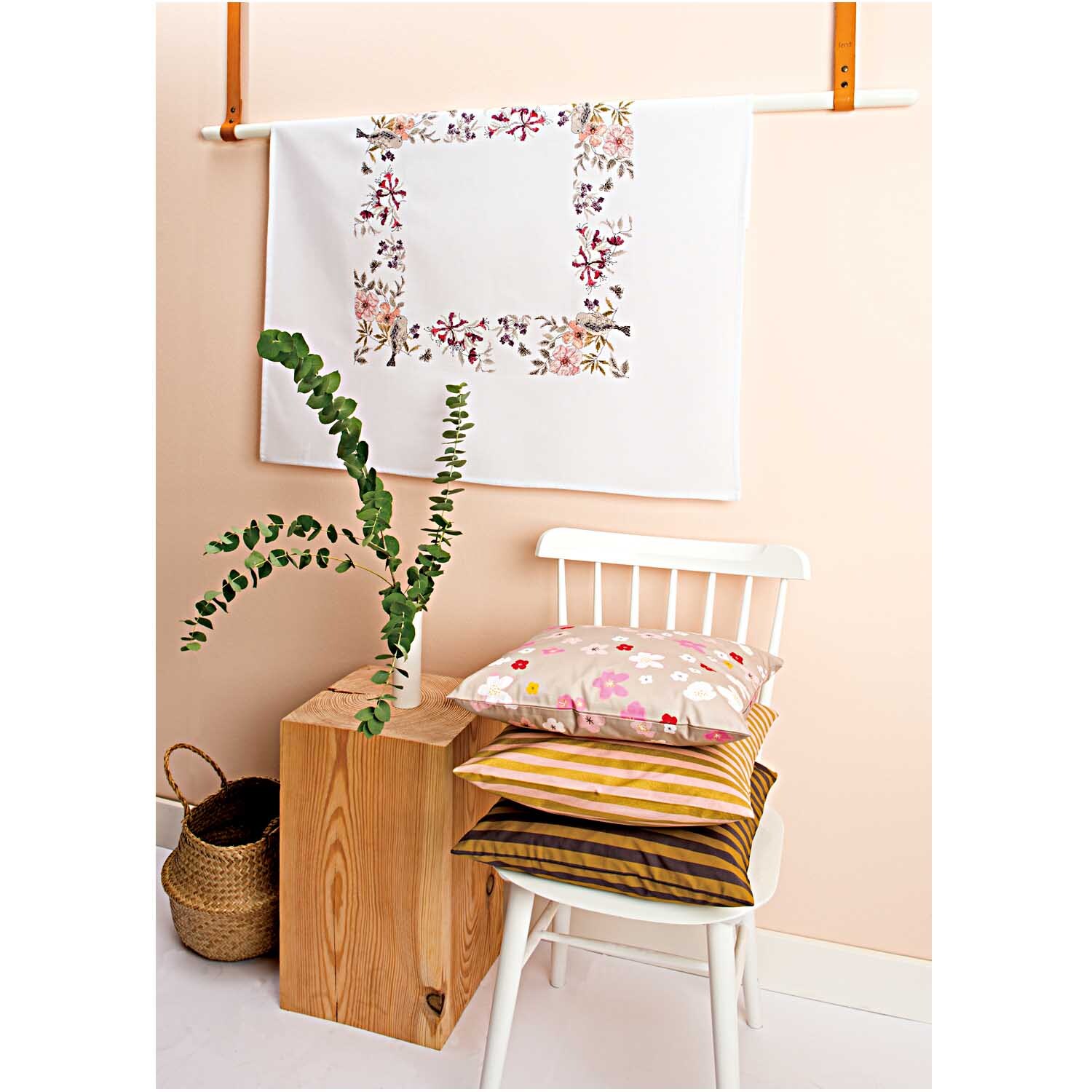Stickpackung Tischdecke Vogel 90x90cm von Rico Design, aufgehängt an einer Wand, umgeben von Pflanzen und Kissen auf einem Stuhl.