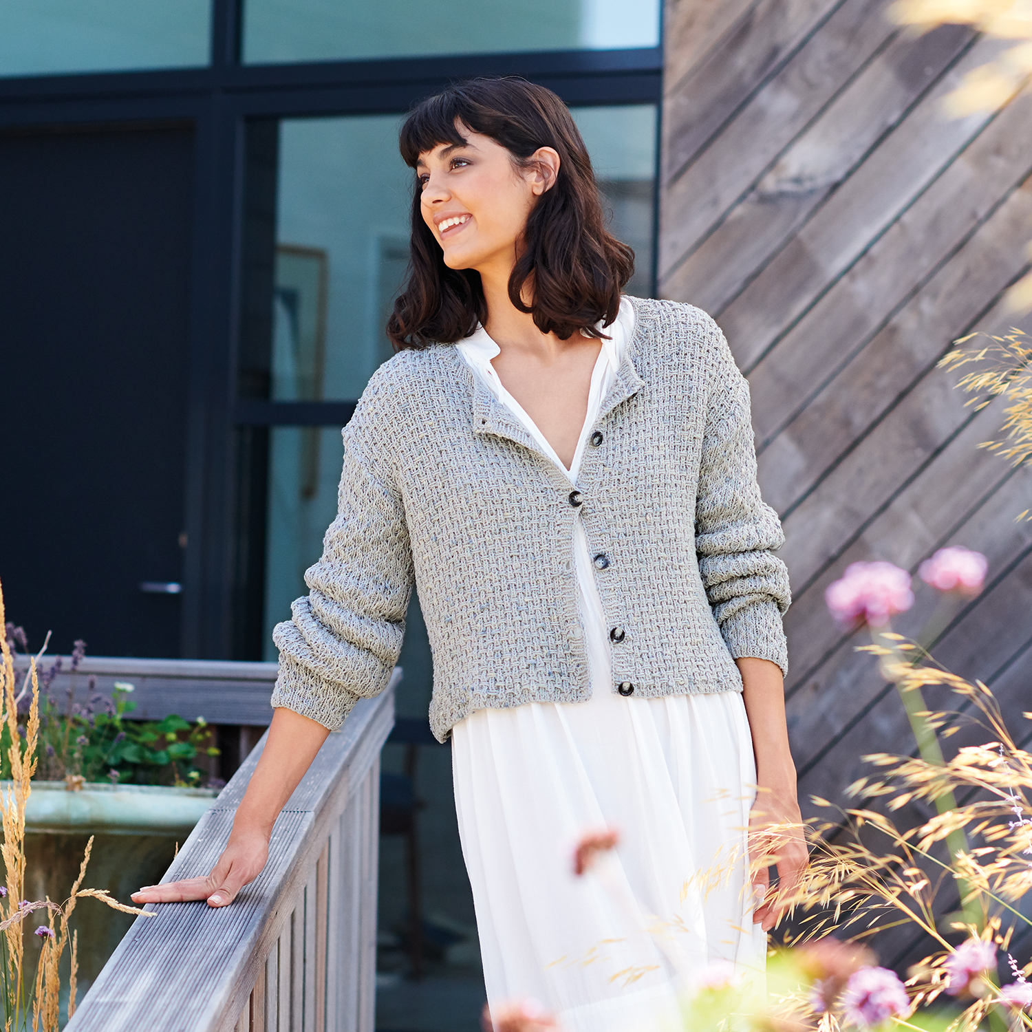 Strickanleitung Strukturmuster-Jacke aus Fashion Cotton Neps dk: Frau in grauer Strickjacke lehnt lächelnd an einem Geländer, umgeben von Blumen.