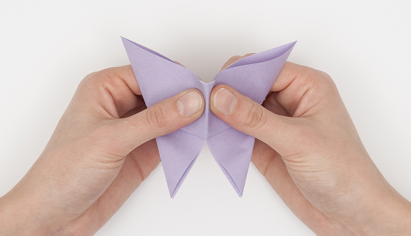 Bastelanleitung Origami-Schmetterlinge: Hände falten einen lila Origami-Schmetterling auf weißem Hintergrund.