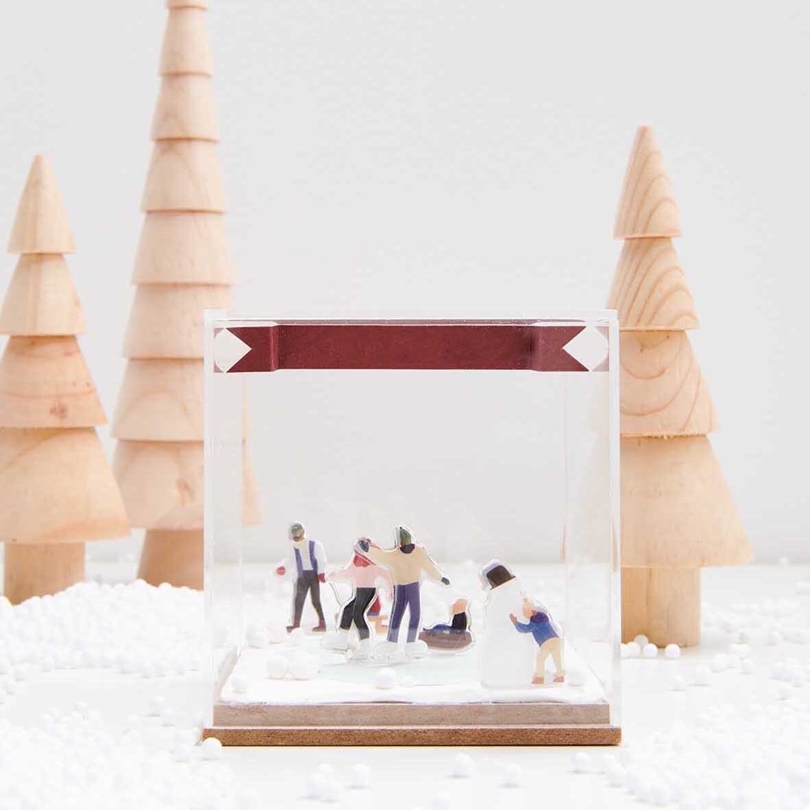 Figurico Gelsticker Schneevergnügen zeigt eine winterliche Szene mit Figuren in einem klaren Würfel, umgeben von Holz-Tannenbäumen.