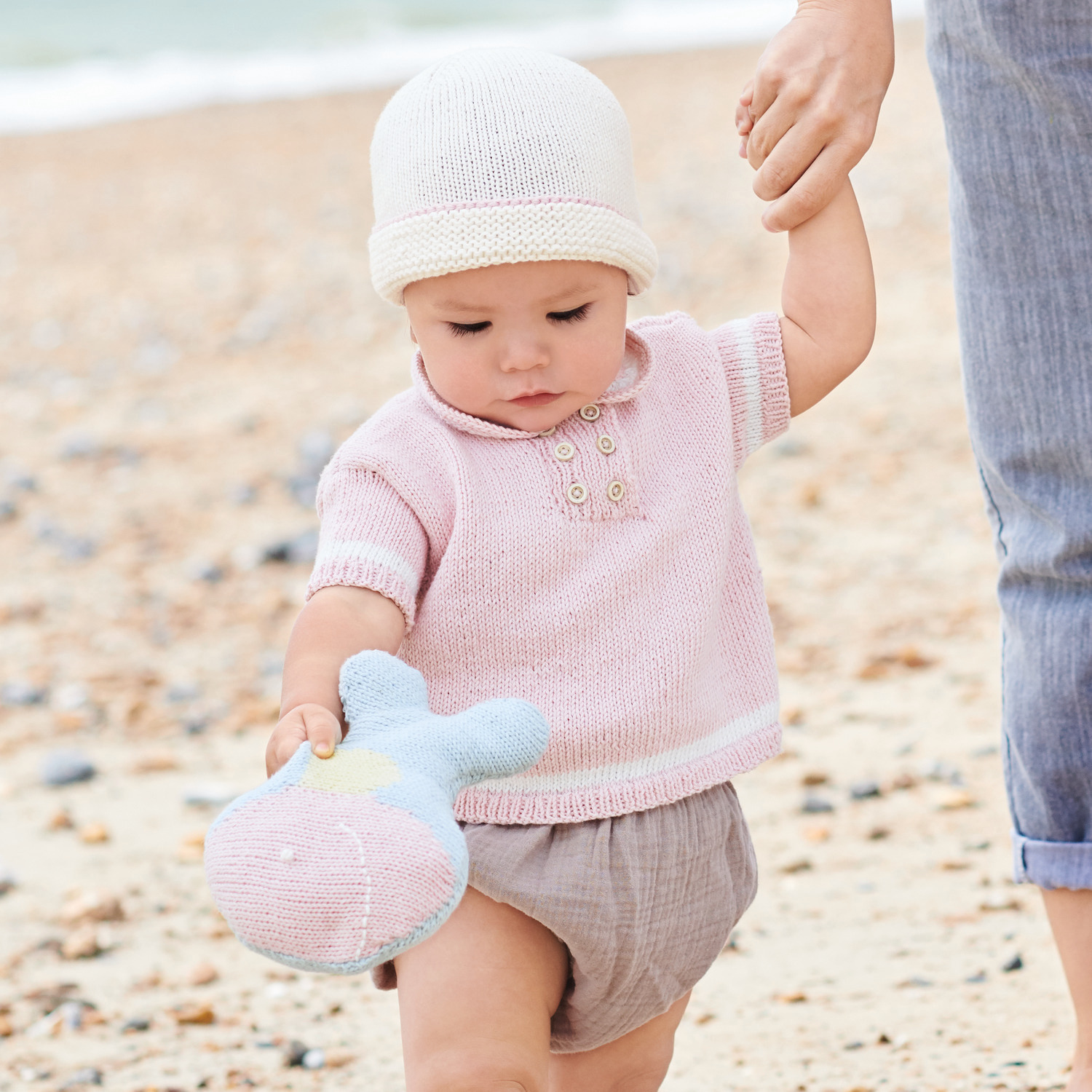Strickset Shirt Modell 16: Baby in rosa Strickshirt und Mütze hält hellblaues Kuscheltier am Strand.