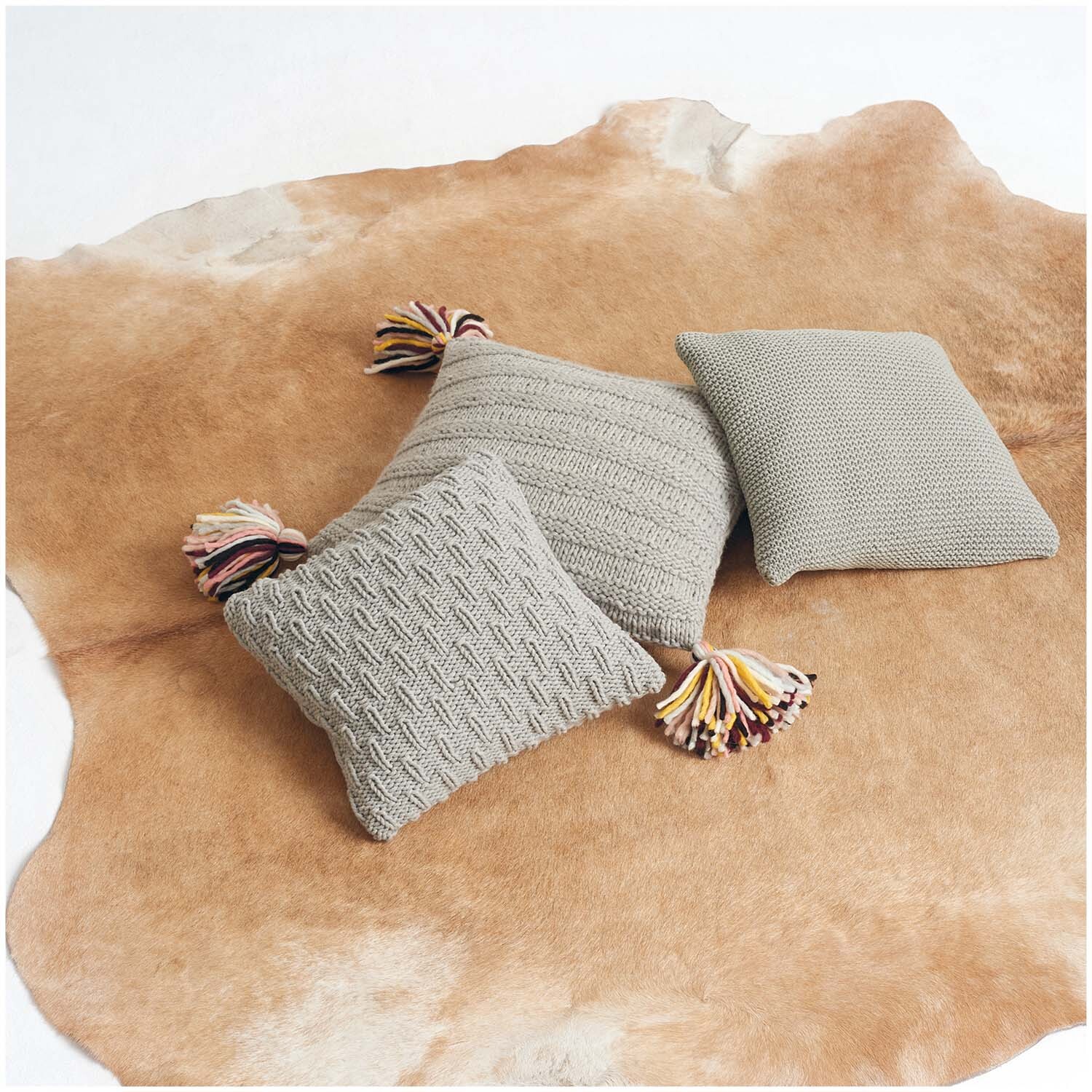 Classik Kissen auf einem hellbraunen Fellteppich, gestrickt mit Wolle Rödel Rico Design, verziert mit bunten Quasten.