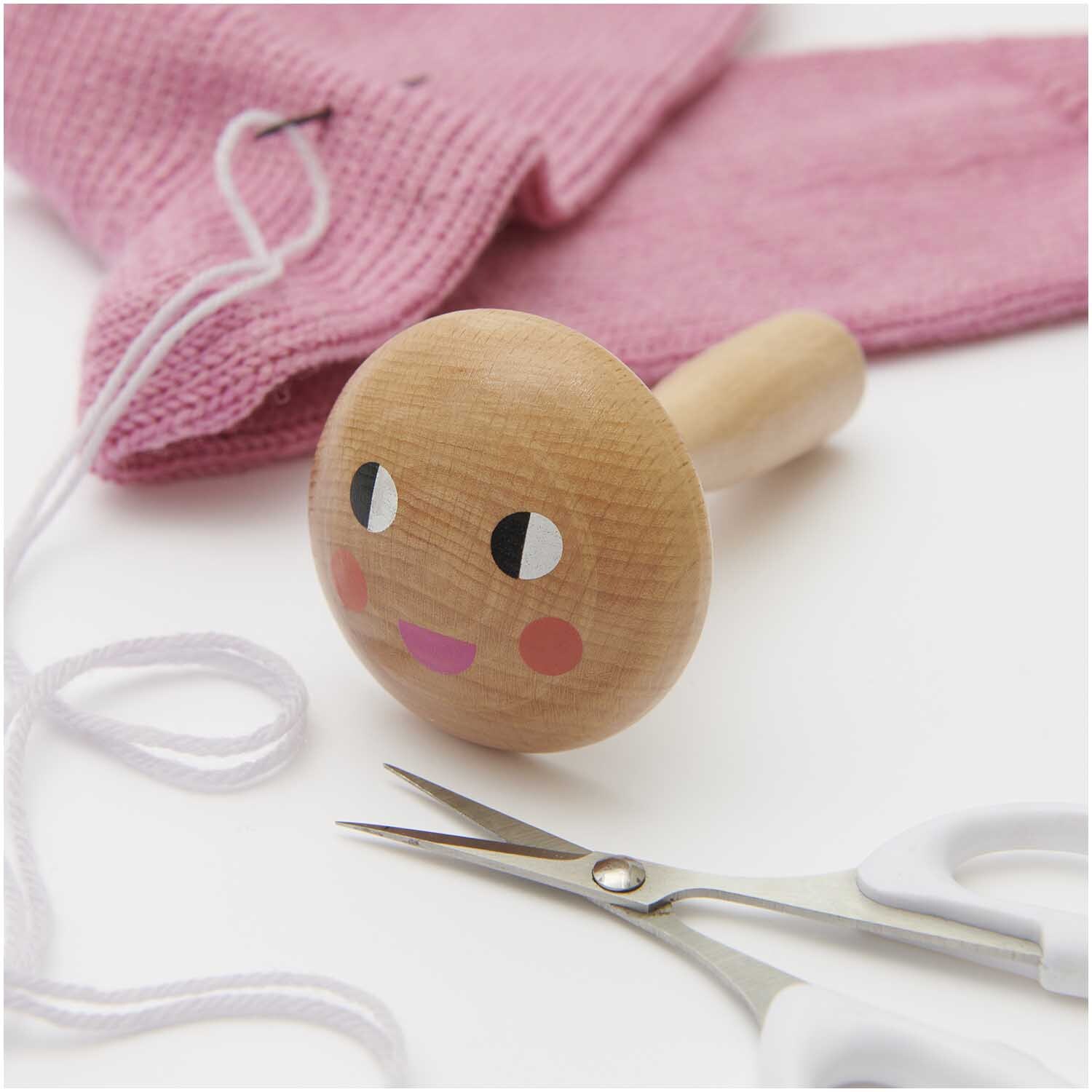 Stopfpilz mit Gesicht aus Holz, umgeben von rosa Strickware, Schere und weißem Faden. Ideal für Handarbeiten.