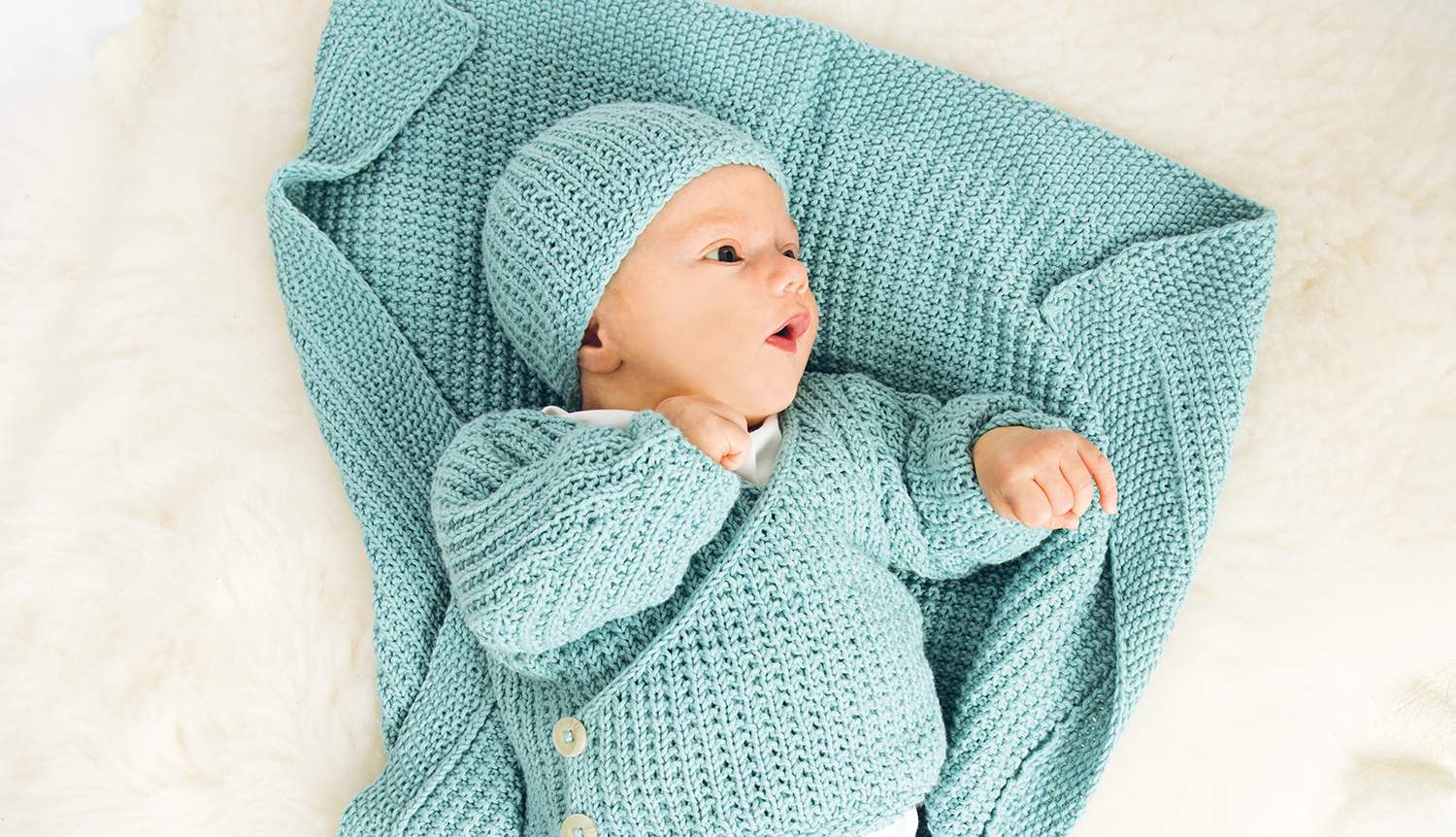 Strickanleitung Baby-Jacke mit Knöpfen: Ein Baby in blauer Strickjacke und Mütze auf weißem Hintergrund.