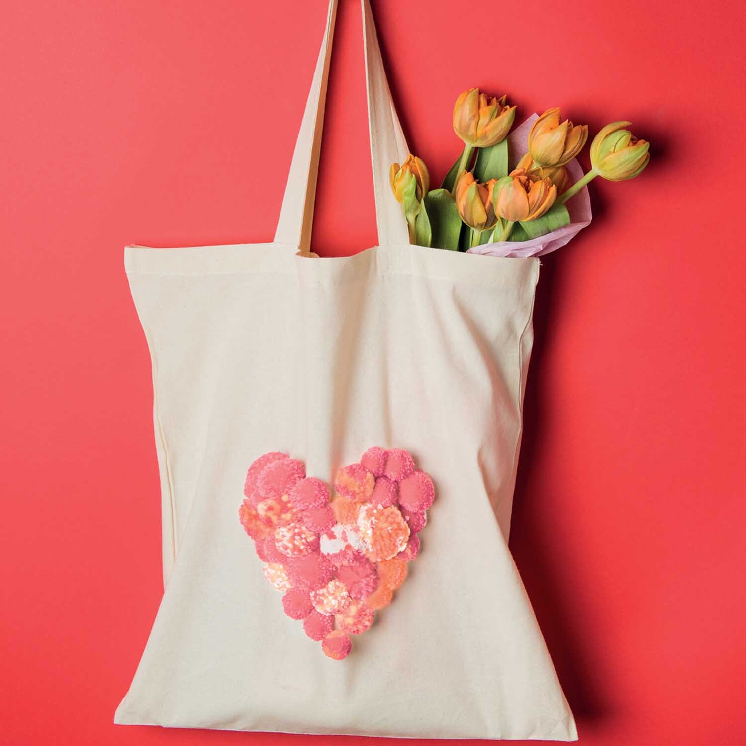 Bastelanleitung Baumwolltasche mit Pompons: Eine Tasche mit Herz aus bunten Pompons, gefüllt mit Tulpen, vor rotem Hintergrund.