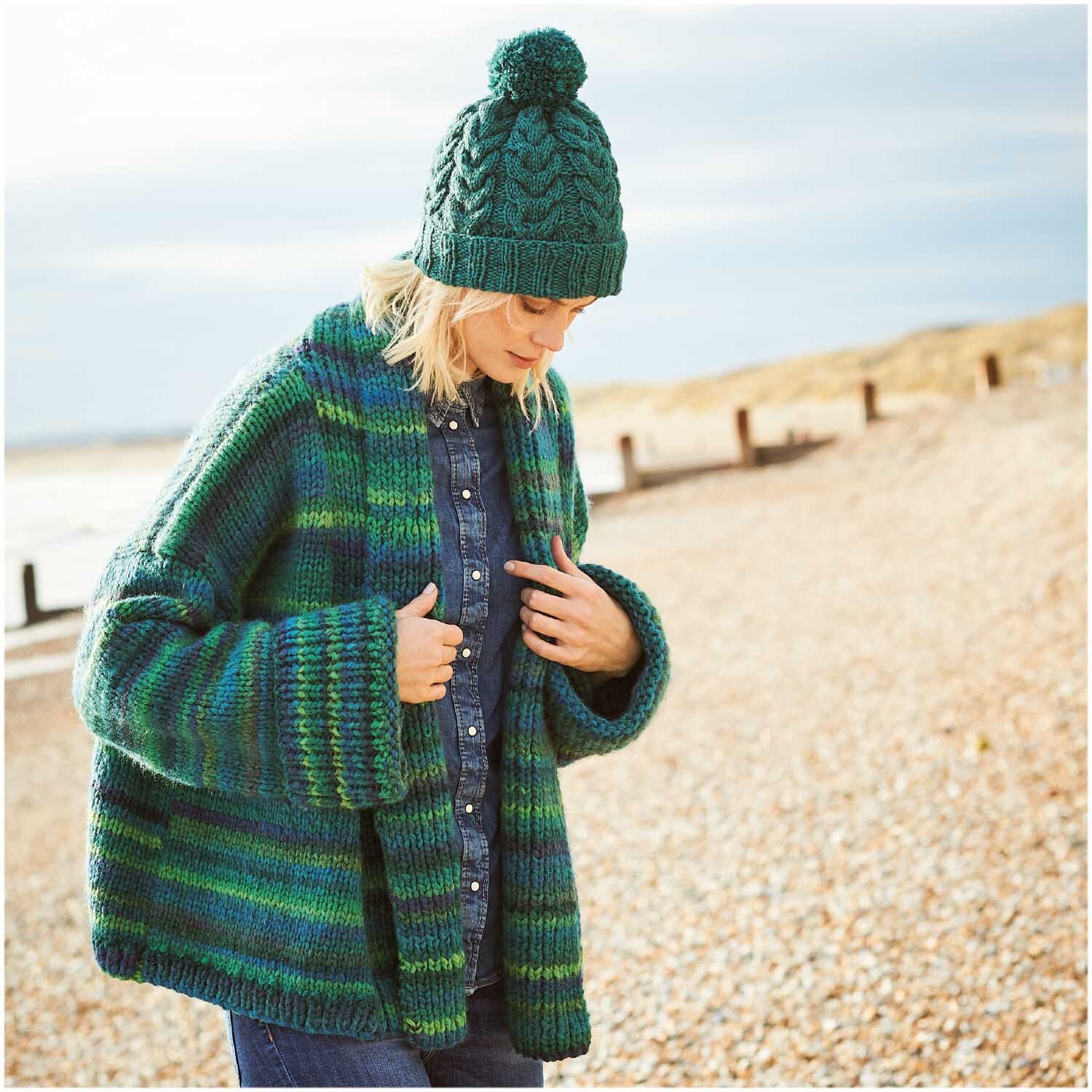 Essentials Alpaca Blend chunky: Frau am Strand in grün-blauem Strickmantel und Mütze von Rico Design.