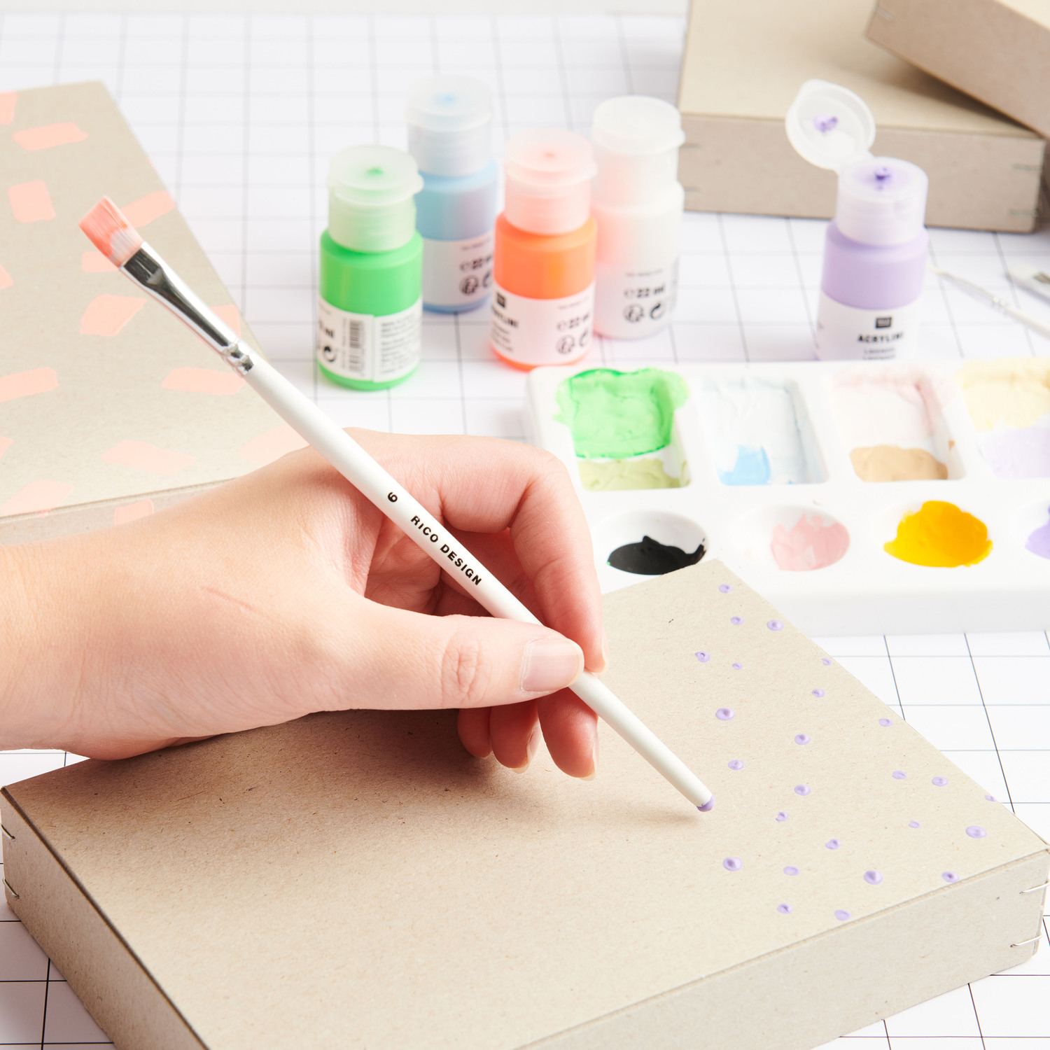 Acrylini Acrylfarben Set Power Colours: Eine Hand malt mit Pinsel auf Karton, umgeben von bunten Farben und Malutensilien.