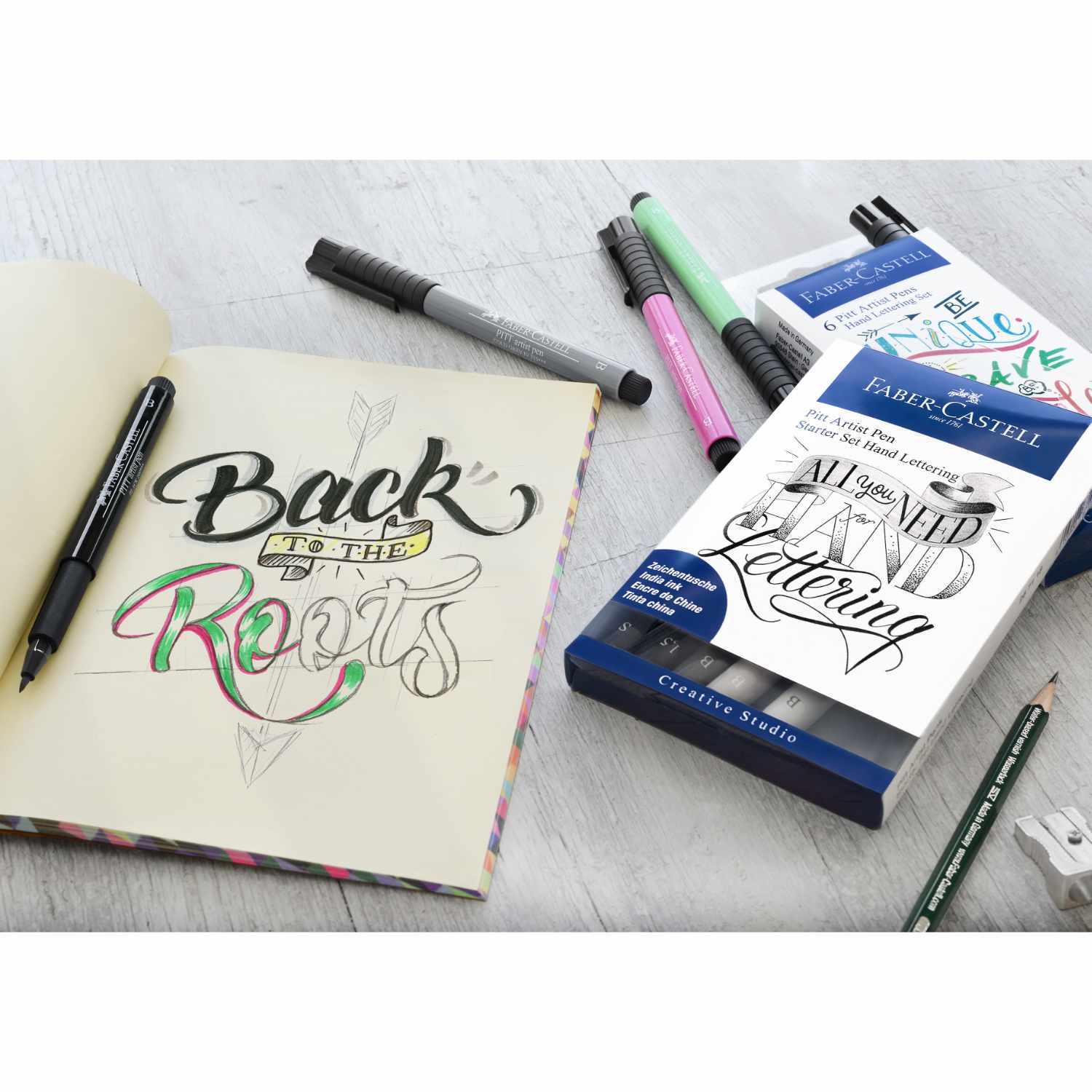 Tuschestift Pitt Artist Pen Handlettering 4 Stück von Faber Castell neben einem Skizzenbuch mit Schriftzug. Ideal für kreative Designs.