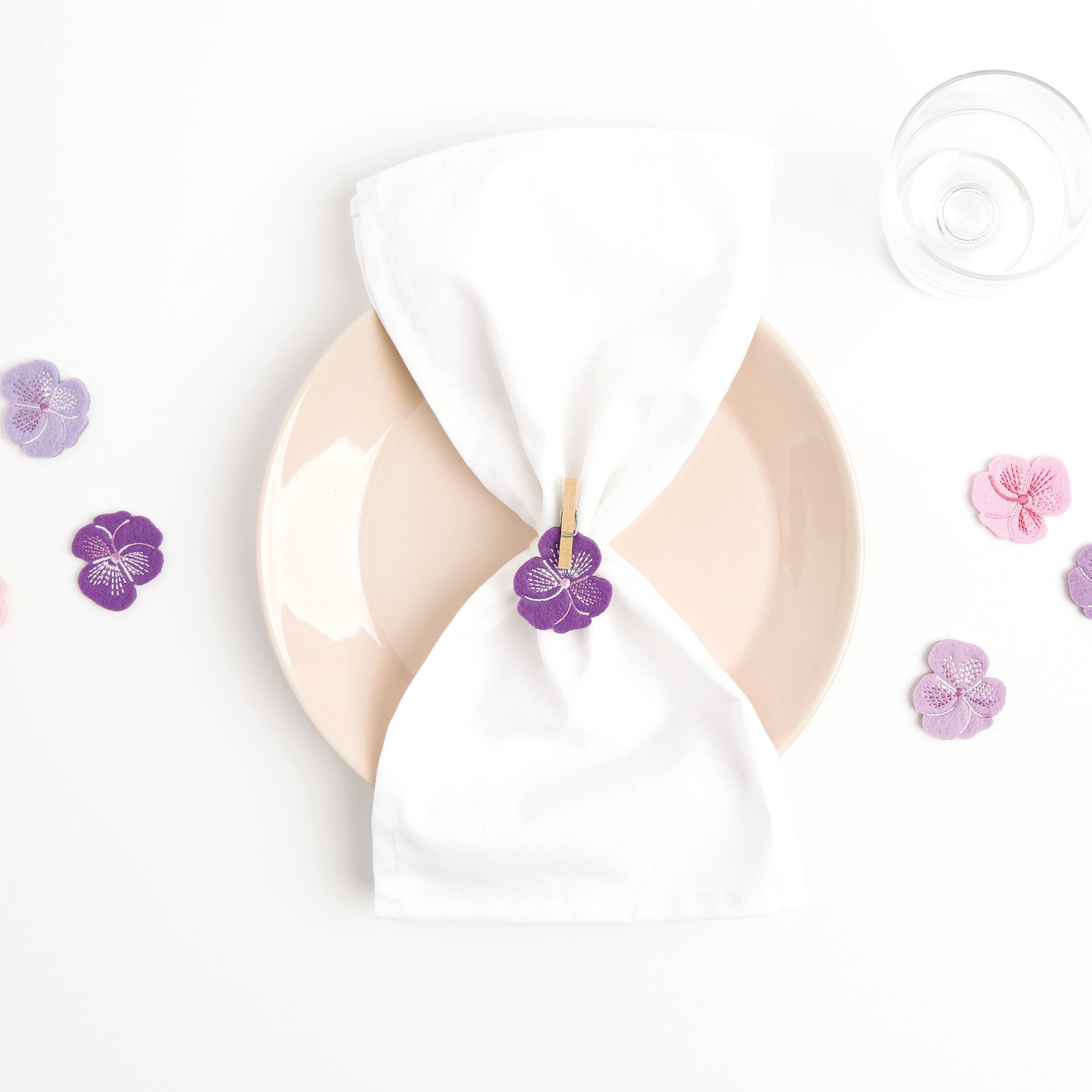 Filzstreu Stiefmütterchen bestickt Rosa, dekorativ auf einem Teller arrangiert, mit Serviette und Glas. Ideal für stilvolle Tischdekoration.