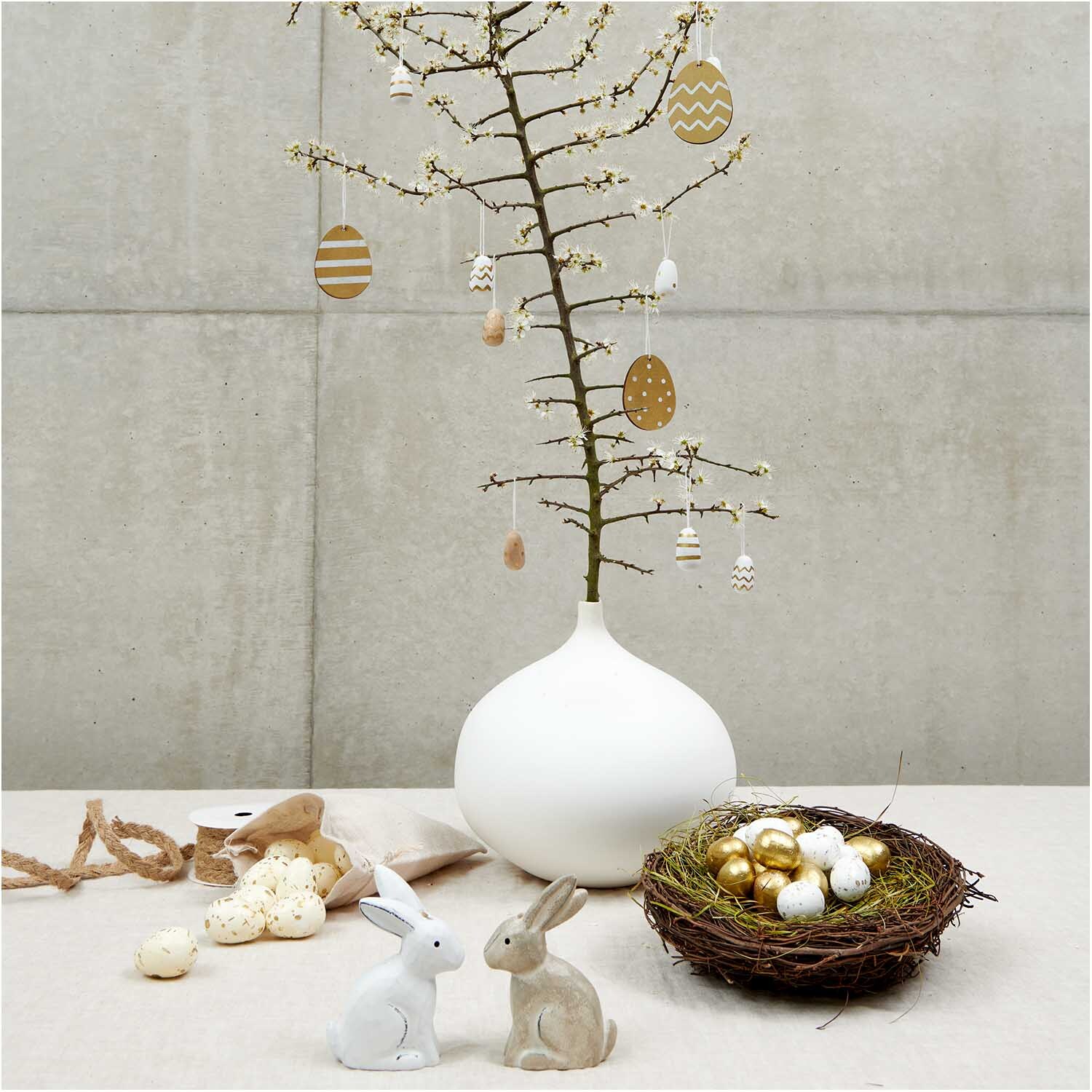 Eier im Netz 4cm 24 Stück: Dekorative Ostereier an einem minimalistischen Baum in einer weißen Vase, umgeben von Hasenfiguren und einem Nest.