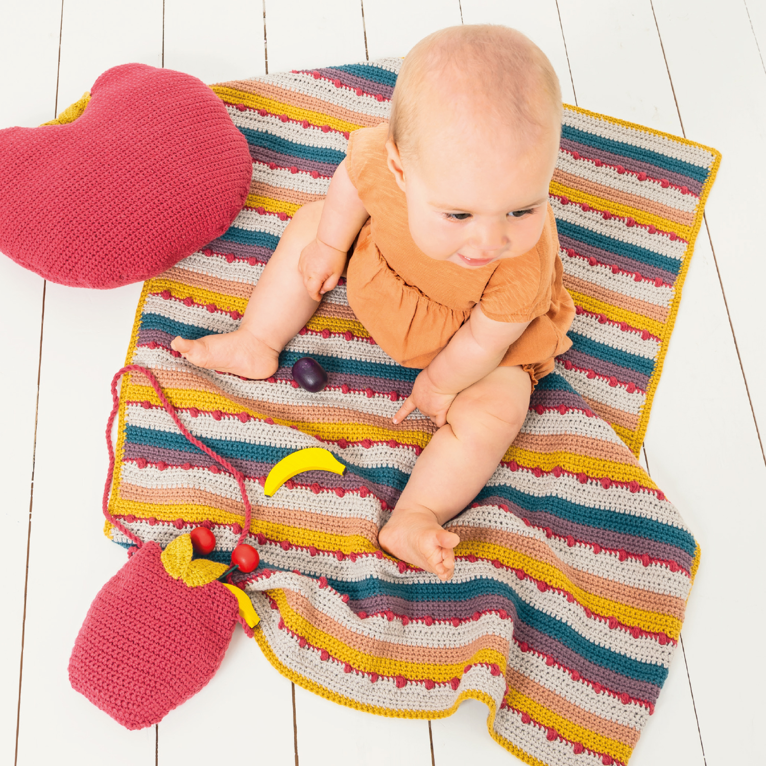 Häkelanleitung Decke aus Baby Cotton Soft DK: Ein Baby sitzt auf einer bunten, gestreiften Decke mit Spielzeug.