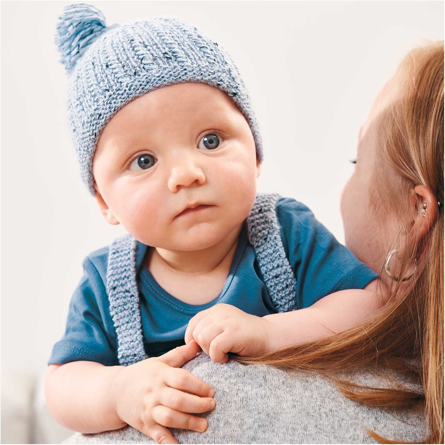 Rico Baby Dream Tweed dk: Ein Baby mit blauer Strickmütze und passendem Outfit auf dem Arm einer Frau.