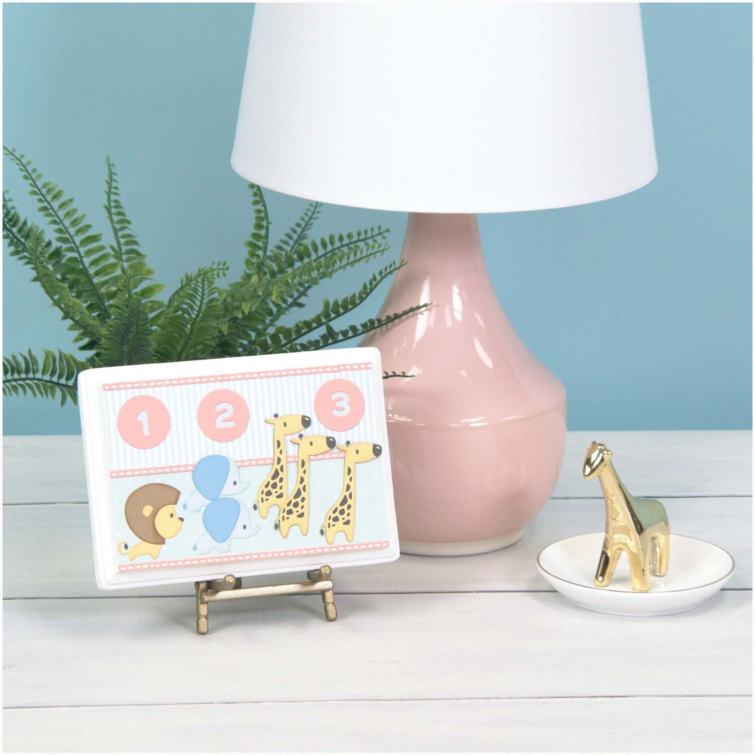 Thinlits Die Set Baby Jungle Animals zeigt eine Karte mit Dschungeltieren neben einer rosa Lampe und einem goldenen Giraffen-Dekor.
