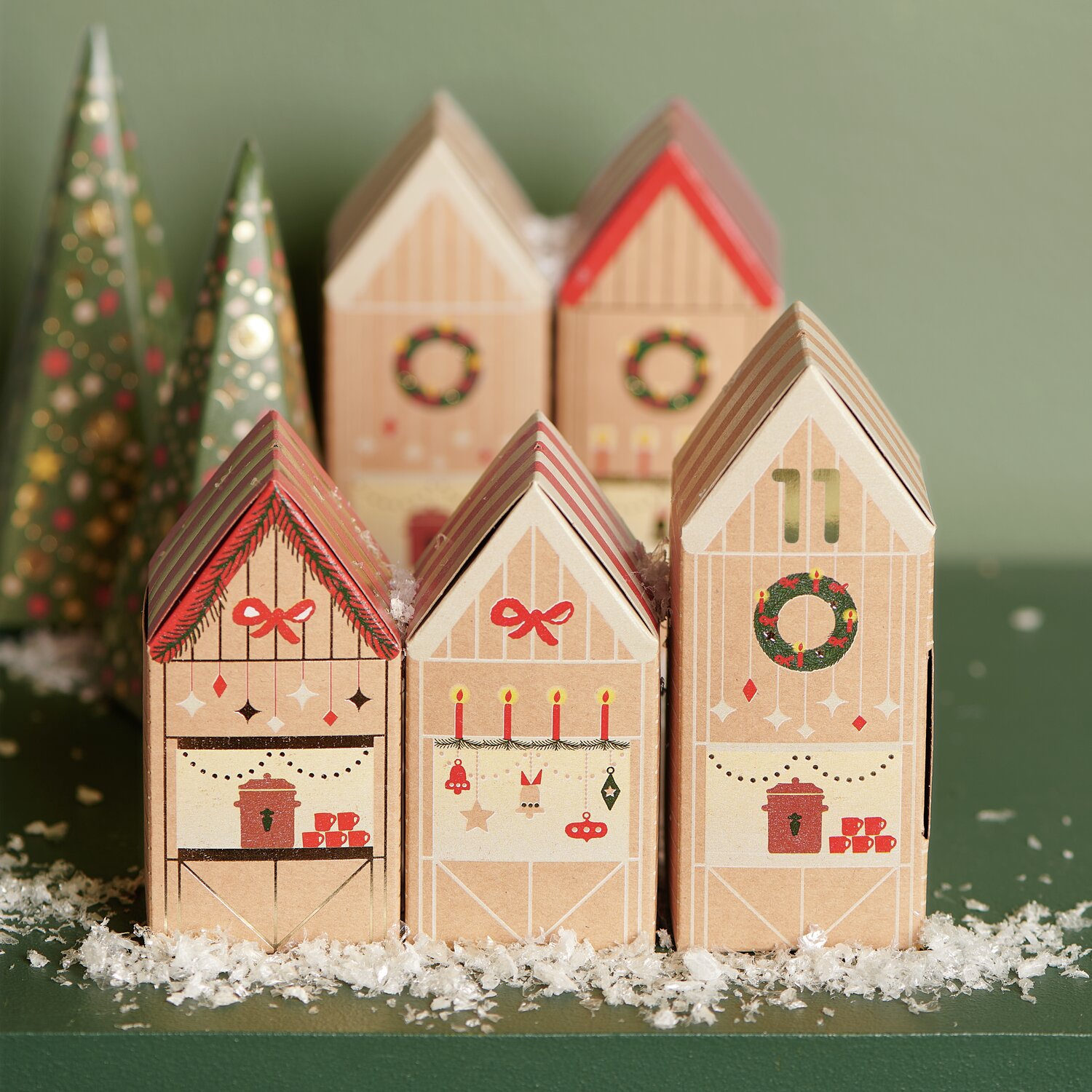 Adventskalender Bastelset mit festlichen Häuschen in Beige und Rot, dekoriert mit Kränzen und Schleifen, auf grünem Hintergrund mit Kunstschnee.
