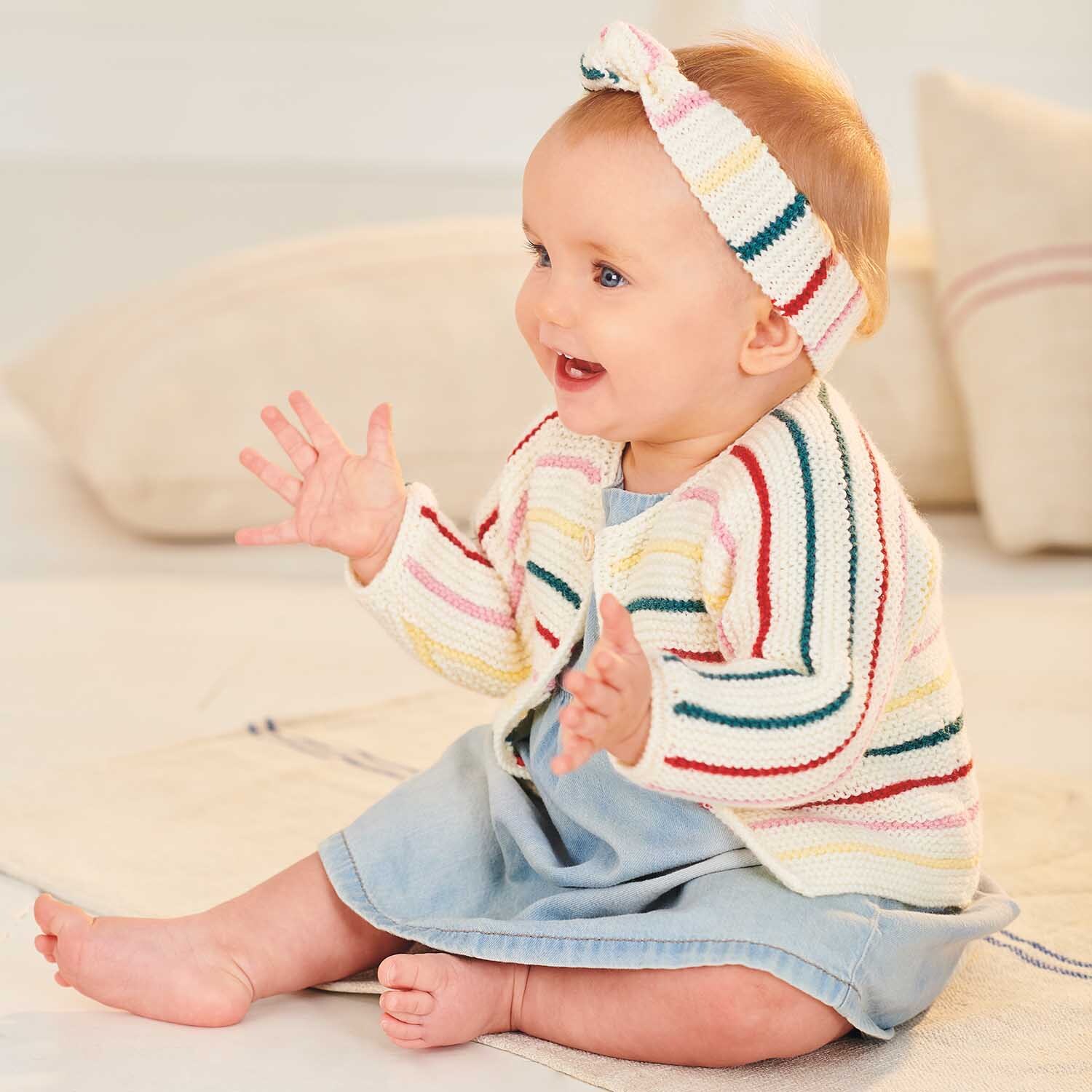 Strickanleitung Stirnband & Jacke aus Rico Baby Classic dk: Baby trägt gestreiftes Set in Pastellfarben, sitzt auf hellem Teppich und lächelt.