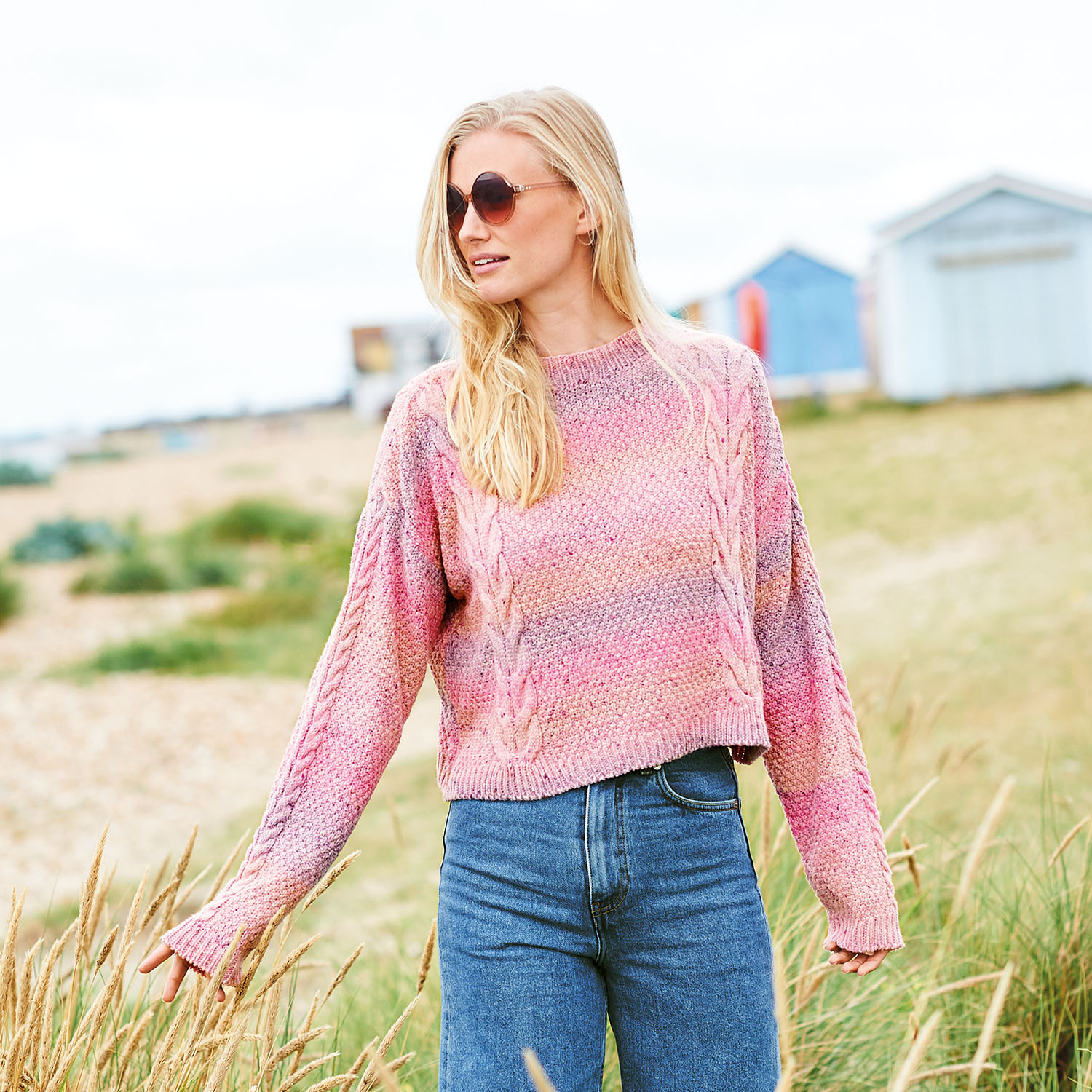 Strickanleitung Pullover aus Fashion Cotton Light & Long Tweed dk: Frau am Strand in rosa Pullover und Jeans, umgeben von Strandhütten und Gras.