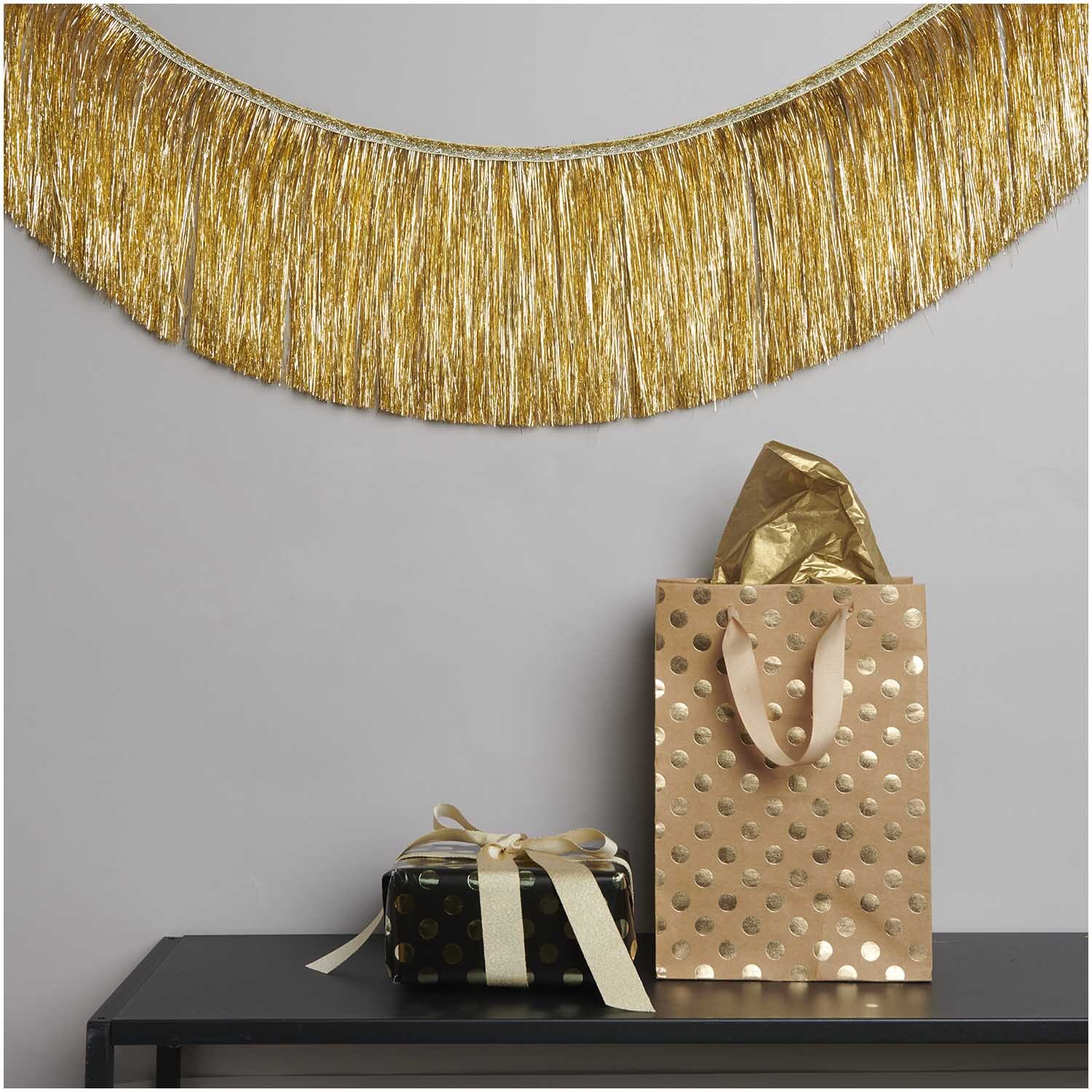 Lametta Girlande gold 3,5m von Rico Design dekoriert eine Wand über einem Tisch mit Geschenken.