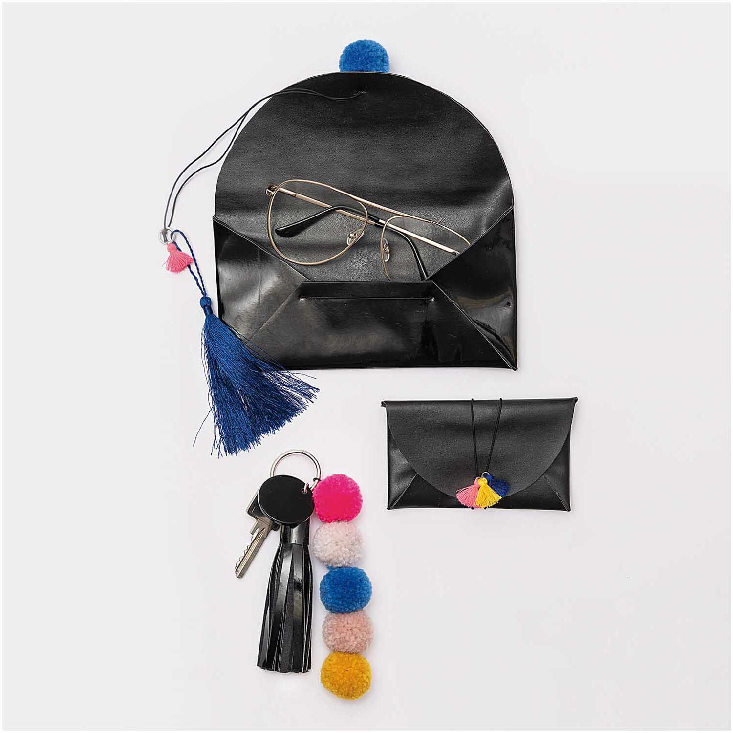 Kunstleder matt 45x100cm: Schwarze Accessoires von Rico Design, darunter eine Brillentasche und Schlüsselanhänger mit bunten Pompons.
