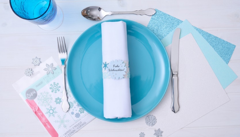 Bastelanleitung Serviettenring mit der Sizzix gestalten: Ein gedeckter Tisch mit blauem Teller, weißer Serviette und dekorativem Serviettenring.