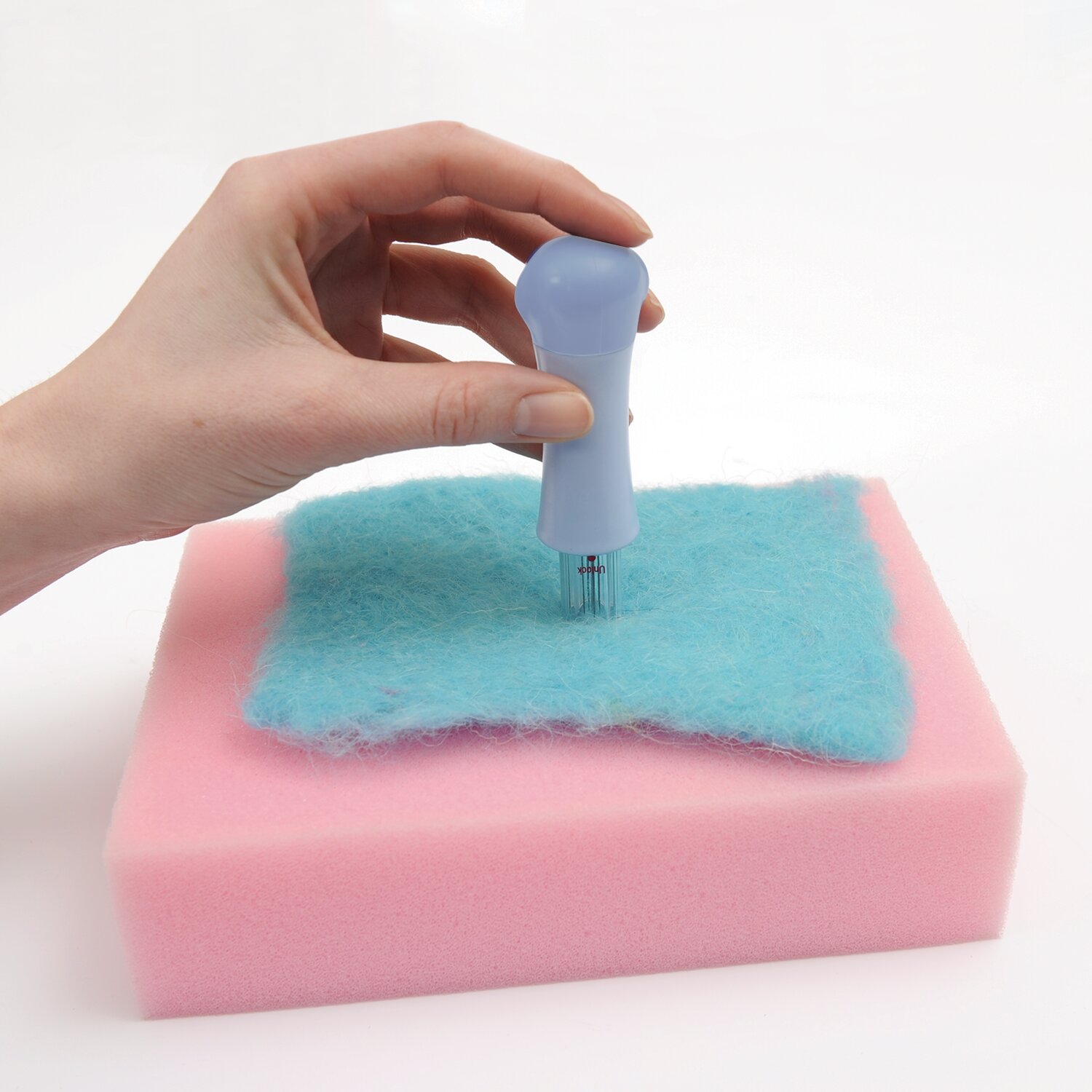 Filznadelhalter inkl. 7 Nadeln von Rico Design, pastellblauer Griff auf rosa Filzunterlage, Hand demonstriert sicheres Trockenfilzen von Wolle
