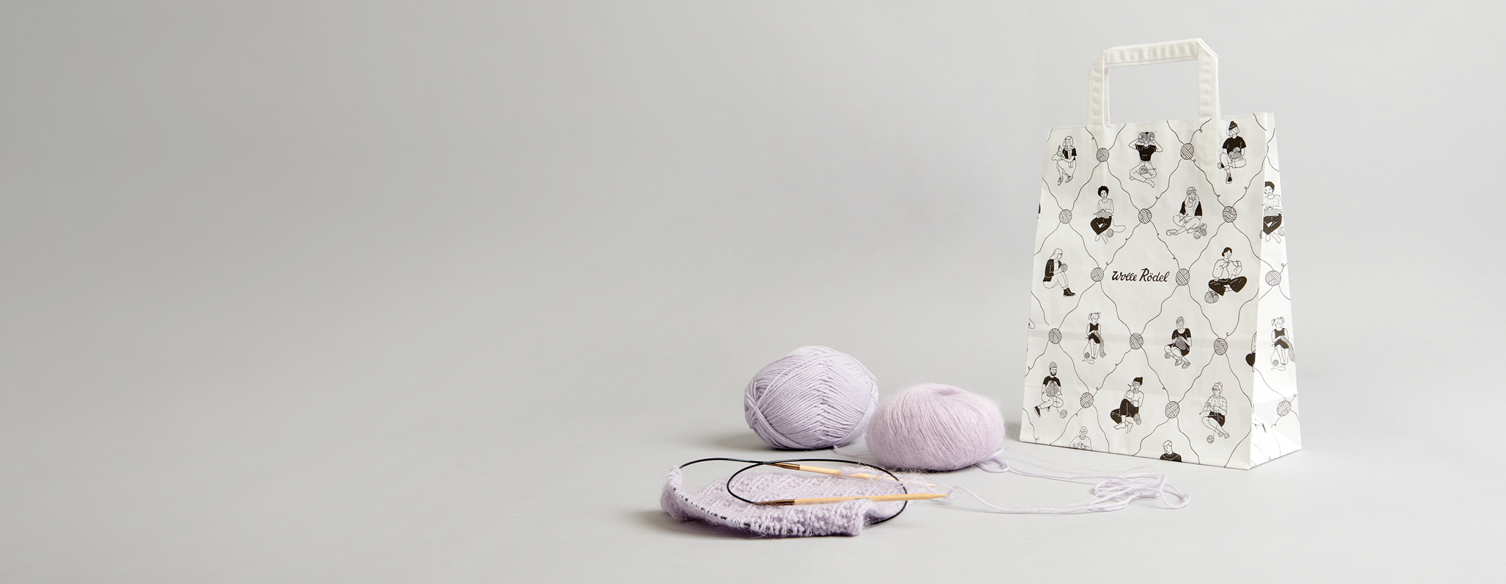 Eine Wolle-Rödel Papiertüte davor ein lila Strickstück, Garn und Rundstricknadeln. Fotosetting graue Wand.