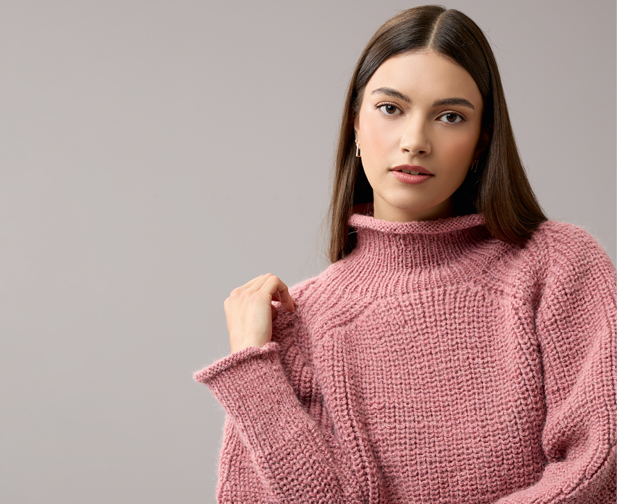 Eine Frau trägt einen Pullover mit hohem Kragen, aus einem altrosa farbigen Garn gestrickt. Fotosetting grauer Hintergrund.