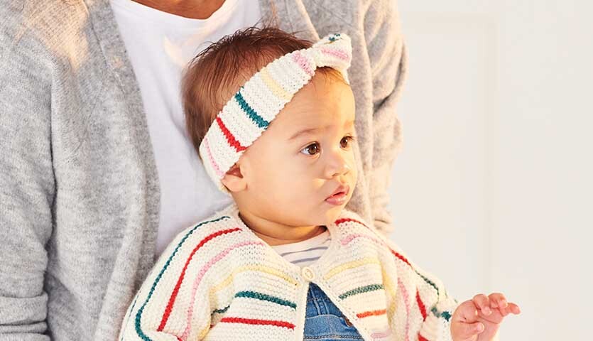 Strickanleitung Stirnband & Jacke aus Rico Baby Classic dk: Baby trägt gestreifte Strickjacke und passendes Stirnband in sanften Farben.