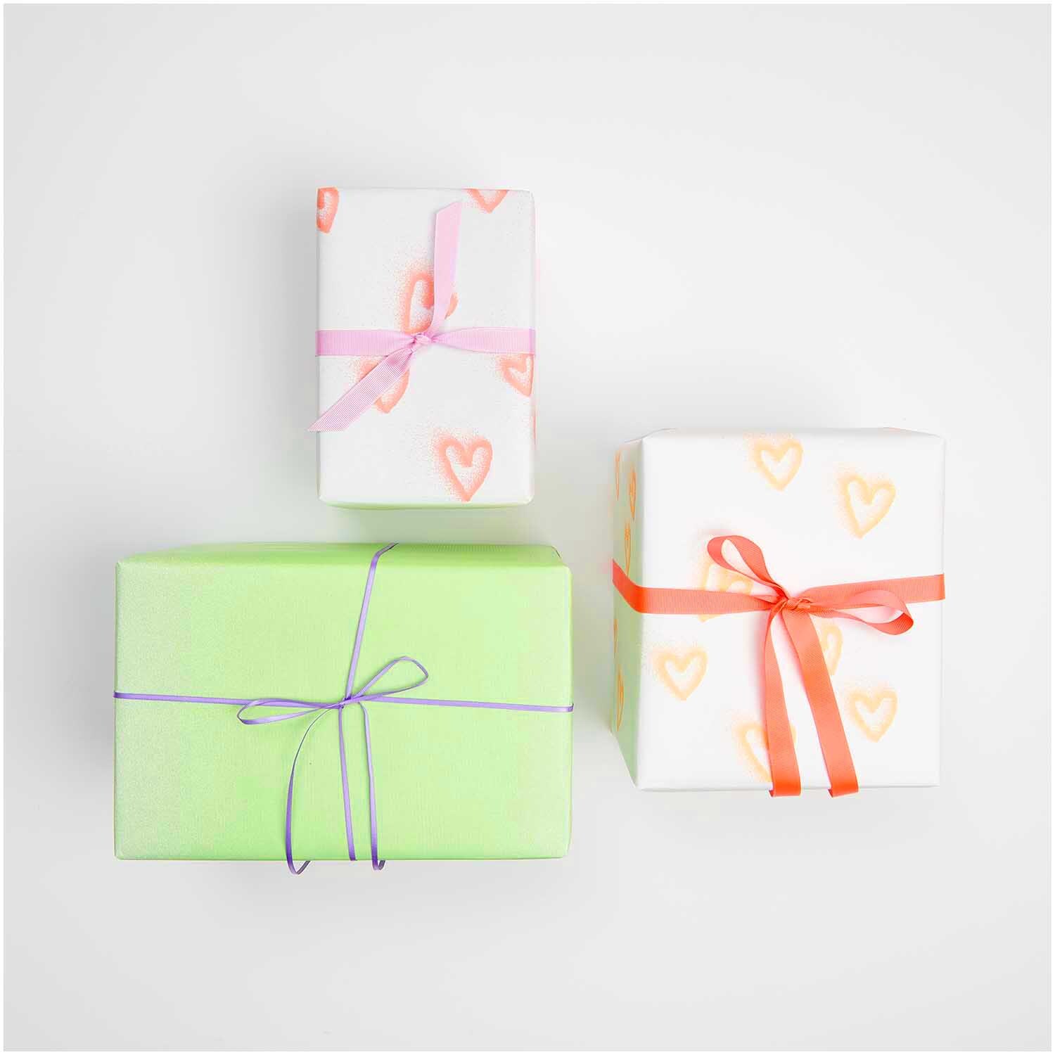Paper Poetry Geschenkpapier zeigt drei Geschenke, zwei mit Herzmuster und eines in Neon-Grün, dekoriert mit bunten Schleifen.