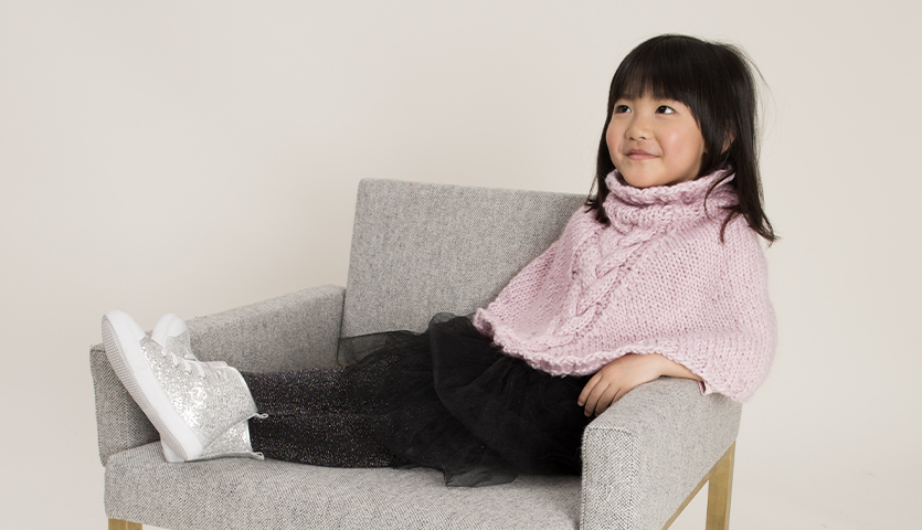 Strickanleitung Kinderponcho aus Glühwürmchen Garn: Mädchen im rosa Poncho auf grauem Sessel.
