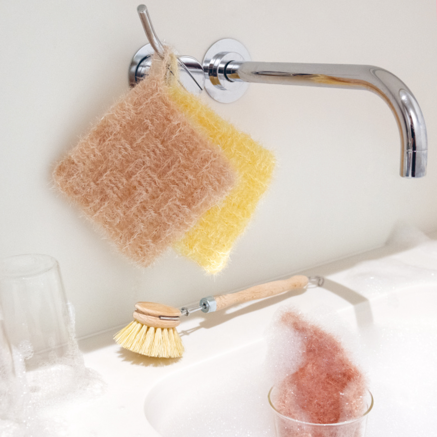 Häkelanleitung Spülschwamm aus Creative Bubble: Zwei Spülschwämme in Beige und Gelb hängen an einem Wasserhahn, daneben eine Spülbürste.